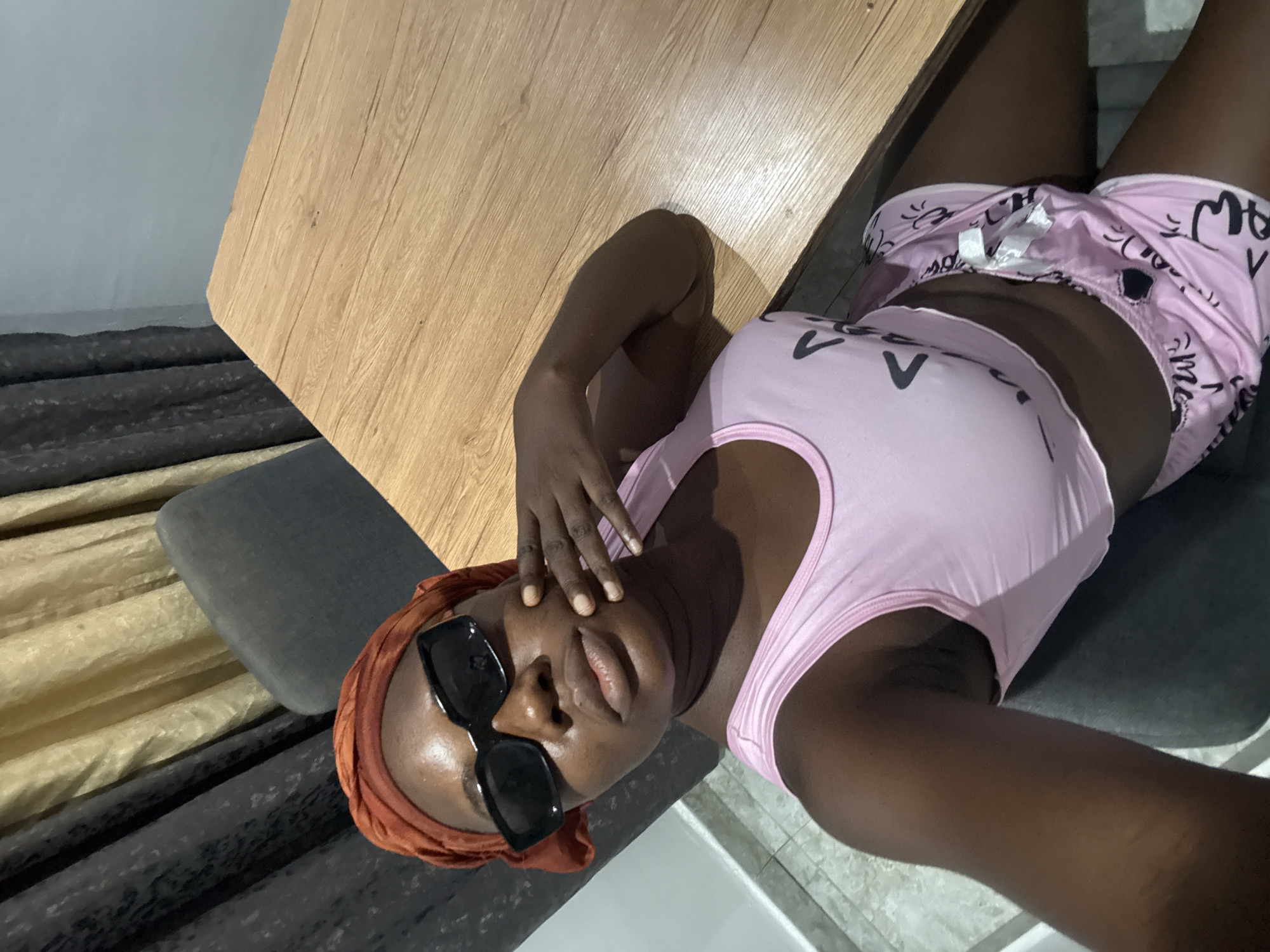 A person with a dark skin tone is captured in a high-angle, rotated selfie, lying on their back indoors. They are wearing a light pink tank top with two black 'V' shapes on the chest, and pink shorts adorned with black patterns and the word 'Meow' visible among other stylized text. Large black sunglasses cover their eyes, and an orange-brown head wrap is worn. One hand is gently placed near their forehead, with fingers visible.
A light wooden surface, possibly a table or desk, diagonally dominates the upper left and central parts of the background. Below this, a dark grey upholstered surface, likely a chair or bed, supports the person. In the background on the left, curtains are visible, featuring sections of beige/cream and dark grey/black fabric. A light grey, possibly tiled, floor can be seen in the lower right corner. The lighting suggests a bright indoor environment during the daytime. The person appears relaxed, looking directly towards the camera. The setting is an indoor space, located in Iddo Tudunwada, Nigeria.