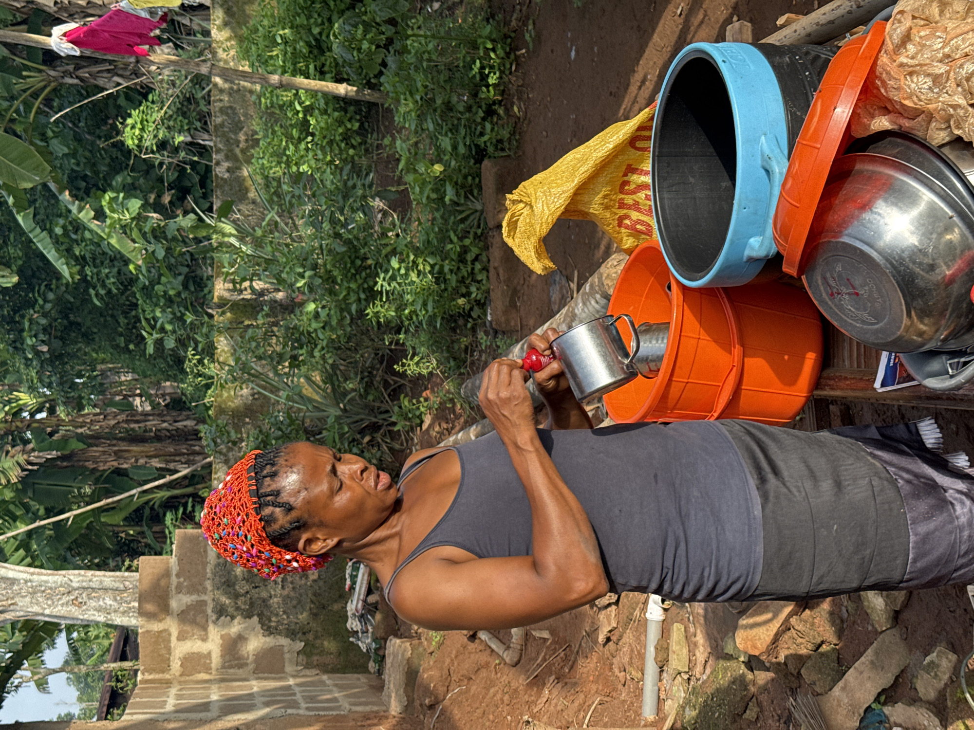 The image, taken in Eziudo, Nigeria, is rotated 90 degrees counter-clockwise and shows a woman engaged in an outdoor task during the daytime. The woman, appearing middle-aged with dark skin, is dressed in a dark grey tank top and a patterned skirt that combines black, grey, and white. Her braided hair is covered by a vibrant orange and multi-colored beaded headband. She holds a silver metal mug in her left hand and a red-handled tool or brush in her right, actively engaged with the mug, possibly cleaning or preparing something.
Around her, a collection of domestic objects is visible. To her right, there is a large black bucket with a distinctive light blue rim and handles, an bright orange bucket, and a reflective silver metal basin. A crumpled yellow plastic bag, clearly displaying the text "BEST O," lies behind these containers. The ground beneath her is an uneven, reddish-brown dirt surface, strewn with rocks and small fragments of debris, including a segment of white pipe.
The background is dominated by lush, dense green foliage, featuring broad leaves characteristic of tropical plants. Portions of concrete block or brick structures are also visible, hinting at a built environment amidst the greenery. In the deeper background, colorful garments, including red and yellow items, hang on what appears to be a clothesline or branch. The bright sunlight and distinct shadows suggest it is a clear day. The woman's expression is focused, indicating concentration on her task.