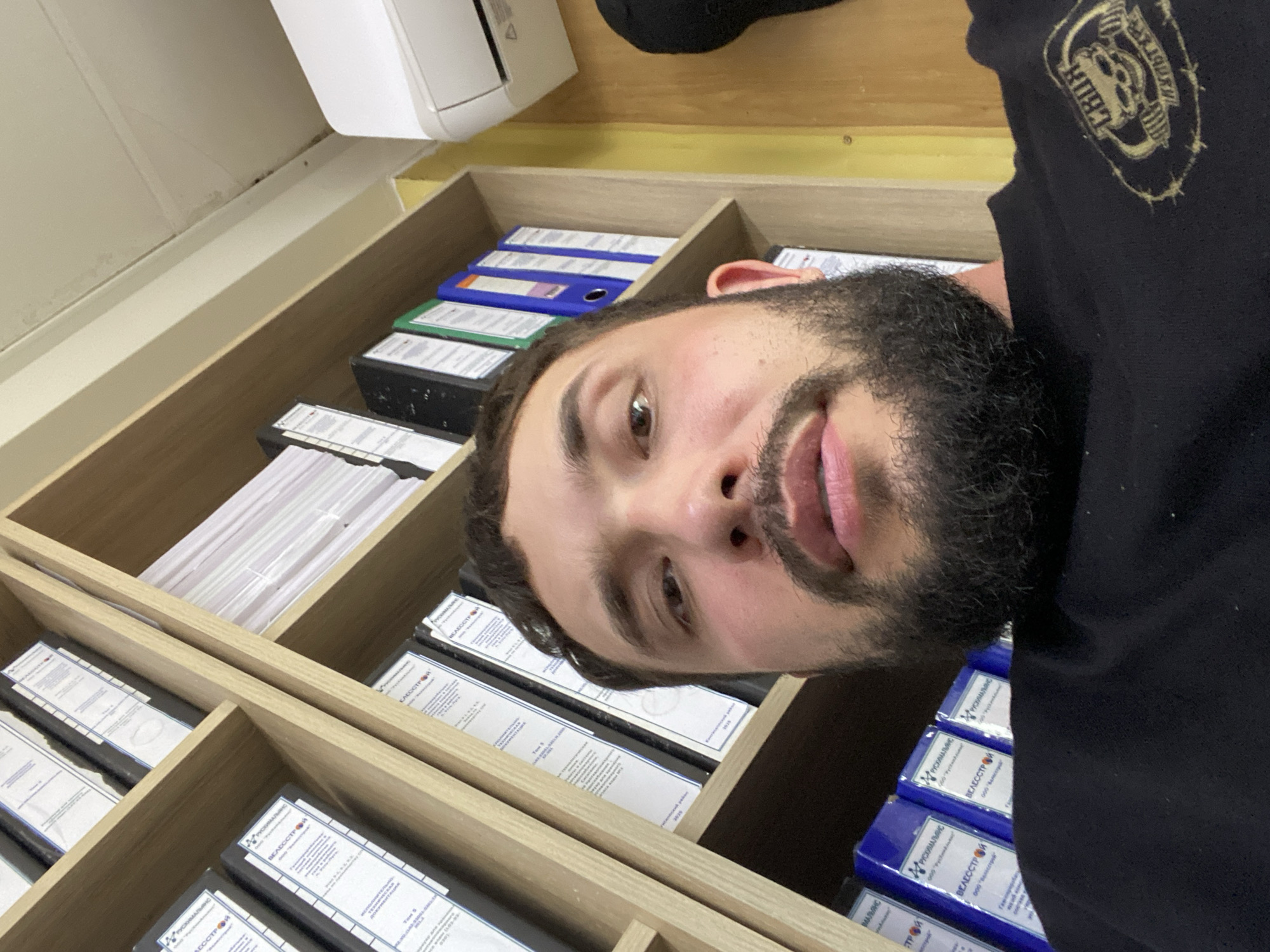 A close-up, low-angle selfie shows a man with dark hair and a beard looking at the camera. He is wearing a black t-shirt with a circular emblem of a skull and weights. He is positioned in front of a wooden bookshelf filled with binders. The background shows an air conditioning unit mounted on a yellow wall, and the ceiling has some signs of wear. The man is identified as being in Luzhitsy, Russia.