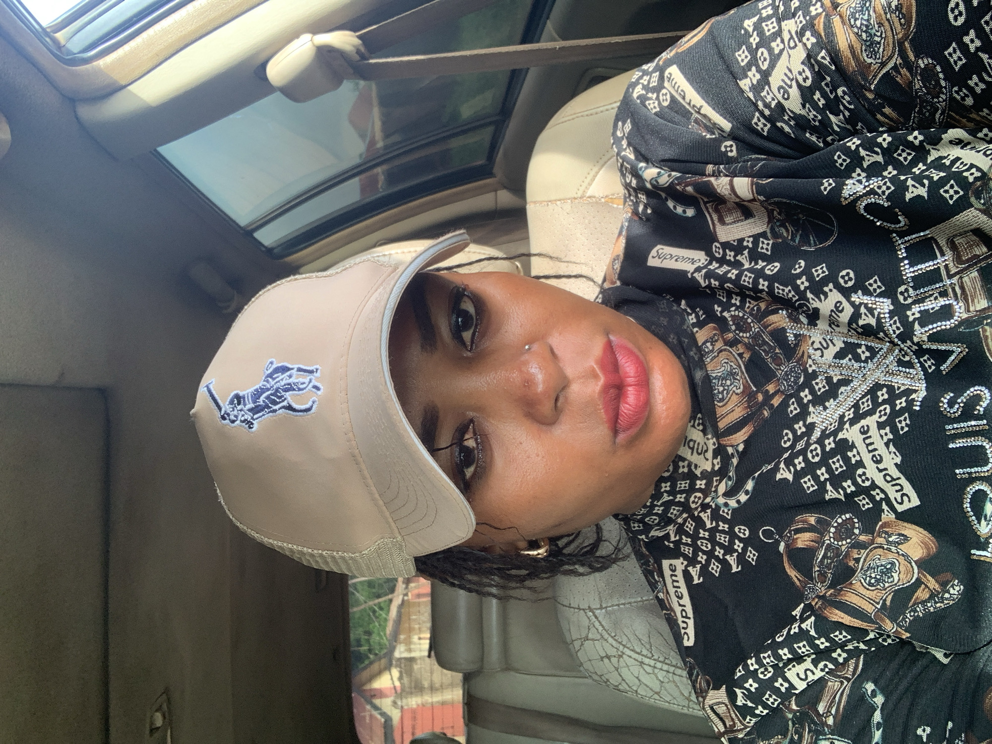 This portrait-oriented image, rotated 90 degrees counter-clockwise, features a woman captured from the chest up, seated inside a car in Sabon Tasha, Nigeria. She looks directly at the camera with a neutral expression. She has a deep skin tone, with a small metallic stud piercing on her left nostril, and her lips are painted with a vibrant red lipstick.
She wears a light beige baseball cap with a dark blue embroidered logo resembling a polo player on a horse. Her top is black and features a rich, busy pattern incorporating elements that evoke Louis Vuitton-style monograms, ornate scrollwork, and the word "Supreme" repeatedly in white rectangular boxes. Parts of the pattern are adorned with small, glittering embellishments, possibly rhinestones or sequins. A gold hoop earring is visible on her left ear.
The background reveals the car's interior, including a light-colored car seat and a sunroof overhead, through which bright daylight is visible, suggesting a clear day. Outside the car windows, hints of trees and buildings are visible, consistent with an urban or residential environment. The overall scene suggests the woman is posing for a casual photo or selfie inside a vehicle during the daytime.