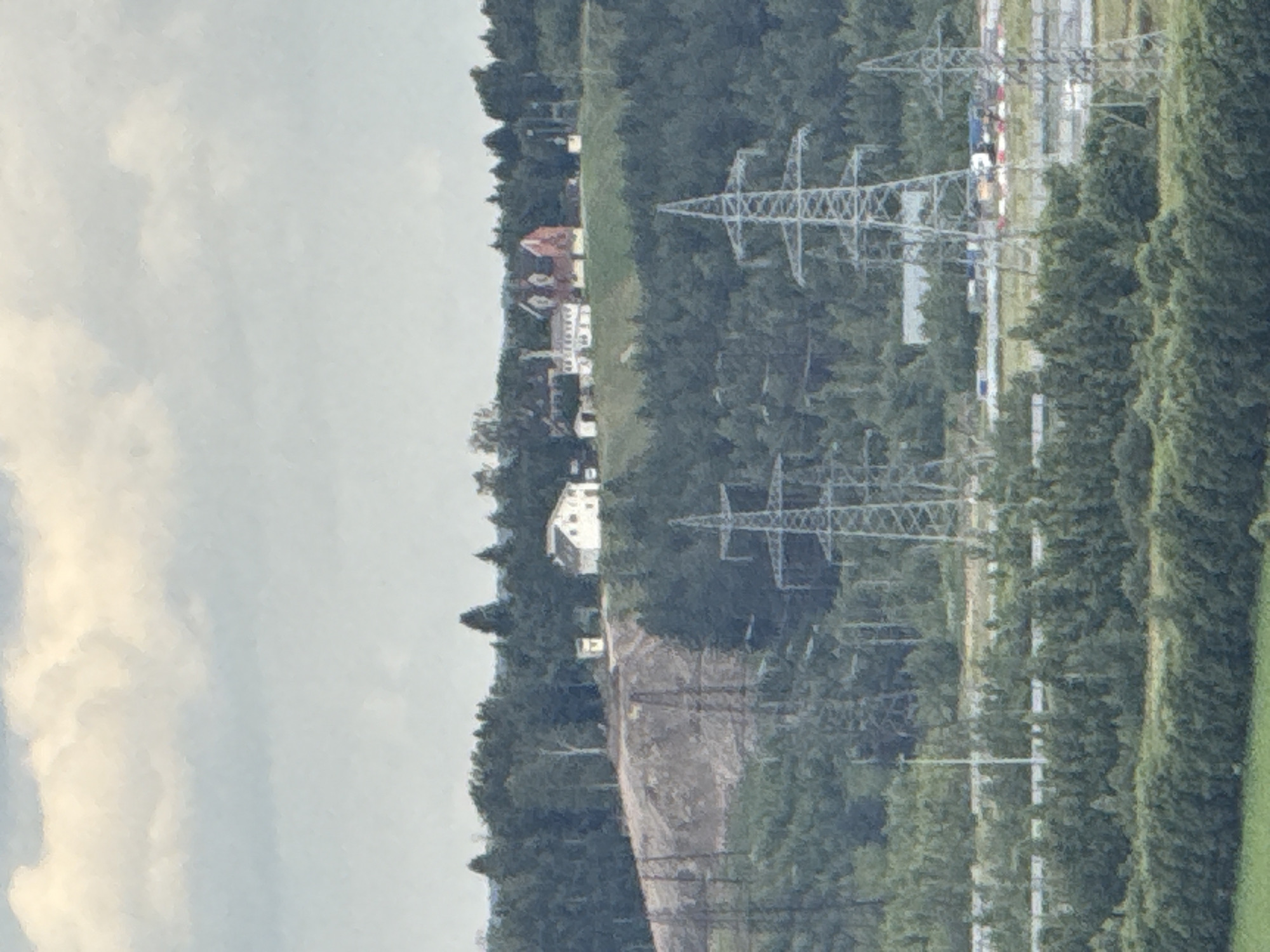 The image depicts a wide shot of a landscape with a focus on industrial and natural elements. In the foreground, several tall electrical transmission towers are visible, supporting high-voltage power lines. Behind these towers, a dense forest covers a hilly terrain. On the crest of one of the hills, a cluster of buildings can be seen, including what appears to be a white house with a red roof and other structures of varying sizes and colors. The background is dominated by a cloudy sky. A road with some vehicles is also partially visible in the mid-ground. This scene is located in the city of Pargolovo, Russia.