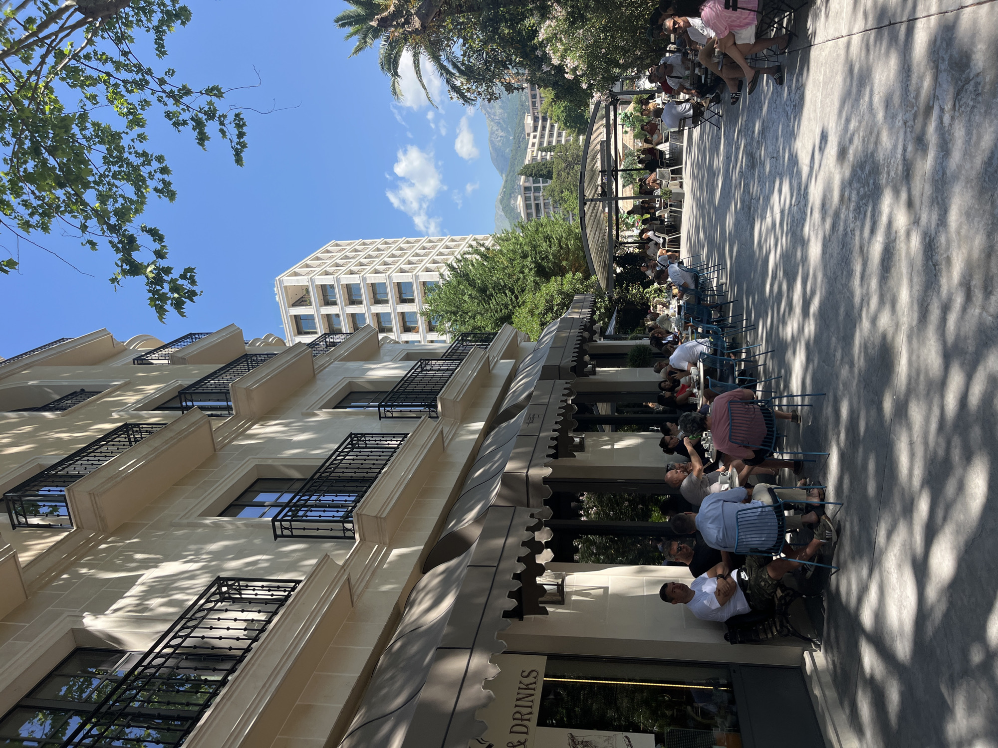 This image captures a vibrant outdoor cafe scene in Budva, Montenegro, under clear blue skies with scattered white clouds and bright sunlight.
In the foreground and mid-ground, a large number of patrons are seated at various outdoor tables, occupying a spacious patio area. The patio features a textured, light grey surface, with distinct dappled shadows from overhead trees. Tables are either square or round, with seating provided by blue metal chairs, black metal chairs, and benches. Most individuals are dressed in casual summer clothing, engaging in conversations, with drinks, likely coffee or other beverages, visible on tables. On the far left, a multi-story building with a light cream facade and multiple windows, each featuring black wrought-iron balconies, forms the backdrop for the initial seating area. Several taupe-colored, scalloped awnings extend from this building, providing shade to tables directly beneath.
In the mid-ground right, a larger, open-air seating area is covered by a glass-roofed pergola structure, under which more tables and patrons are visible. Beyond this, a lush green environment of trees and shrubs, including a prominent palm tree on the right, separates the immediate cafe area from the distant background.
In the background, a tall, modern, white-colored building with numerous windows rises on the left. Further back, beyond the greenery, a series of white and light-colored buildings are visible against rolling green mountains under the bright sky. Tree branches with green leaves are also visible in the upper right quadrant of the image, further contributing to the natural shade. The scene depicts a leisurely, social outdoor gathering.