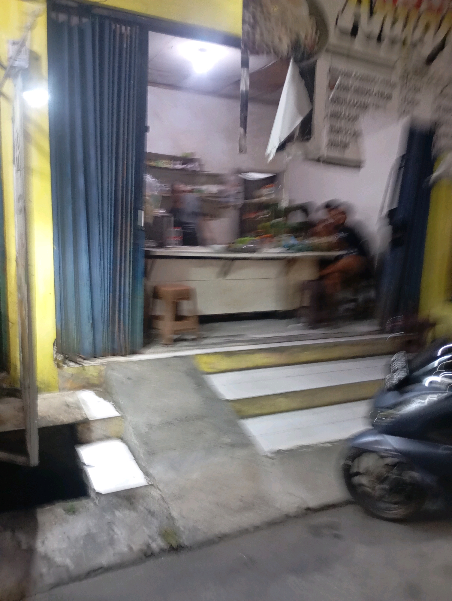 The image captures a street-level view of a storefront in Babelan, Indonesia, likely at night. The storefront is accessed by a short set of stairs and a ramp. A blue, collapsible metal door is partially open, revealing the interior. Inside, a person is seated at a counter, and shelves stocked with items are visible. To the right of the entrance, a motorcycle is parked. The scene is dimly lit, with artificial lighting illuminating the storefront and the immediate outdoor area. The yellow building facade is prominent on the left side of the frame.