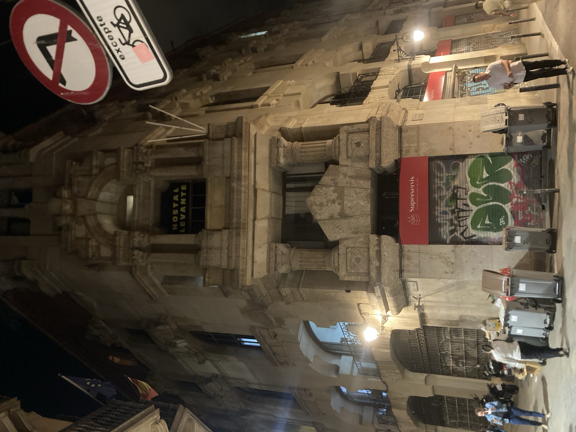 This nighttime image depicts the corner of a multi-story, classically styled stone building in Barcelona, Spain. The structure features ornate stone carvings, pillars with capitals, and arched windows. At an upper level, a yellow sign reads "HOSTAL LEVANTE." On a balcony to the left, a European Union flag and a Spanish flag are displayed.
At ground level, the building features storefronts covered by closed metal roller shutters. Red signage above these shutters identifies "Superservis Supermercats Des de 1945." The lower part of the building facade and the metal shutters are covered in green and red graffiti. A row of five grey wheeled waste bins with brown lids is situated along the sidewalk in front of the building.
On the left side of the street, two women are walking; one wears a denim jacket and dark pants, the other a white top and dark pants. Two motorbikes are parked behind them. On the right side, a man in a white short-sleeved shirt and dark pants walks towards the foreground. Another individual is partially visible further down the street on the right.
Overhead, two traffic signs are visible: a circular red sign with a diagonal black bar, indicating a prohibition, and a rectangular white sign below it with the text "excepte" and a bicycle icon, indicating an exception for bicycles. Street lamps illuminate the scene, casting light and shadows.
The scene is located at the intersection of Baixada de Sant Miquel and other streets in the Gothic Quarter of Barcelona, specifically outside Hostal Levante at Baixada de Sant Miquel, 2, 08002 Barcelona, Spain.
