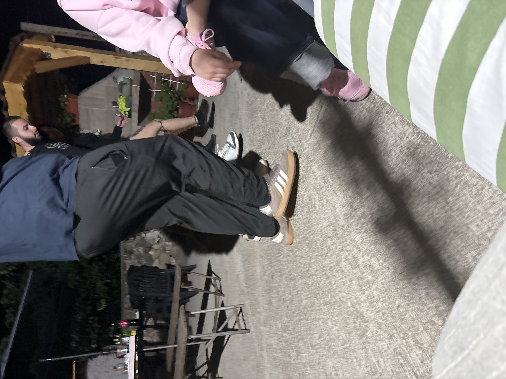The image depicts an outdoor night scene in the city of Krum, Bulgaria, featuring several individuals and a casual gathering. The ground is a light grey, textured surface, likely concrete or paving, illuminated by artificial light sources that cast noticeable shadows.
In the center, a male individual is captured in mid-motion, possibly walking or dancing. He wears a dark blue long-sleeved top and dark grey or black track pants with a zippered pocket on the left thigh. His footwear consists of brown low-top sneakers with white stripes and a gum sole.
To the right of this central figure, partially obscured, stands another male with a beard and short dark hair. He wears a dark long-sleeved top, and his visible right leg exhibits a tattoo near the ankle. He is wearing white sneakers with black accents. Behind him, a wooden pergola or outdoor structure is visible, along with potted plants on a ledge and below.
On the far right, a person (gender unidentifiable due to cropping) is seated, wearing a pink long-sleeved top and dark blue pants. Their hands are clasped, holding a soft, pink, cylindrical or bundled object tied with a pink ribbon. Pink shoes are visible on their feet. A green and white striped cushion or pillow is partially visible in the bottom right corner, suggesting seating.
On the left side of the frame, a rustic-style wooden table with metal folding legs holds various bottles, including clear, green, and a red-labeled bottle (resembling a Coca-Cola product), along with several cups. Behind this table, another similar table is visible, surrounded by dark, folded outdoor chairs. Only the lower legs and feet of a person in dark pants and dark shoes are visible near these tables, suggesting another individual is present, likely seated or standing near the table. Dark foliage or trees are present in the distant background, indicating an outdoor environment.