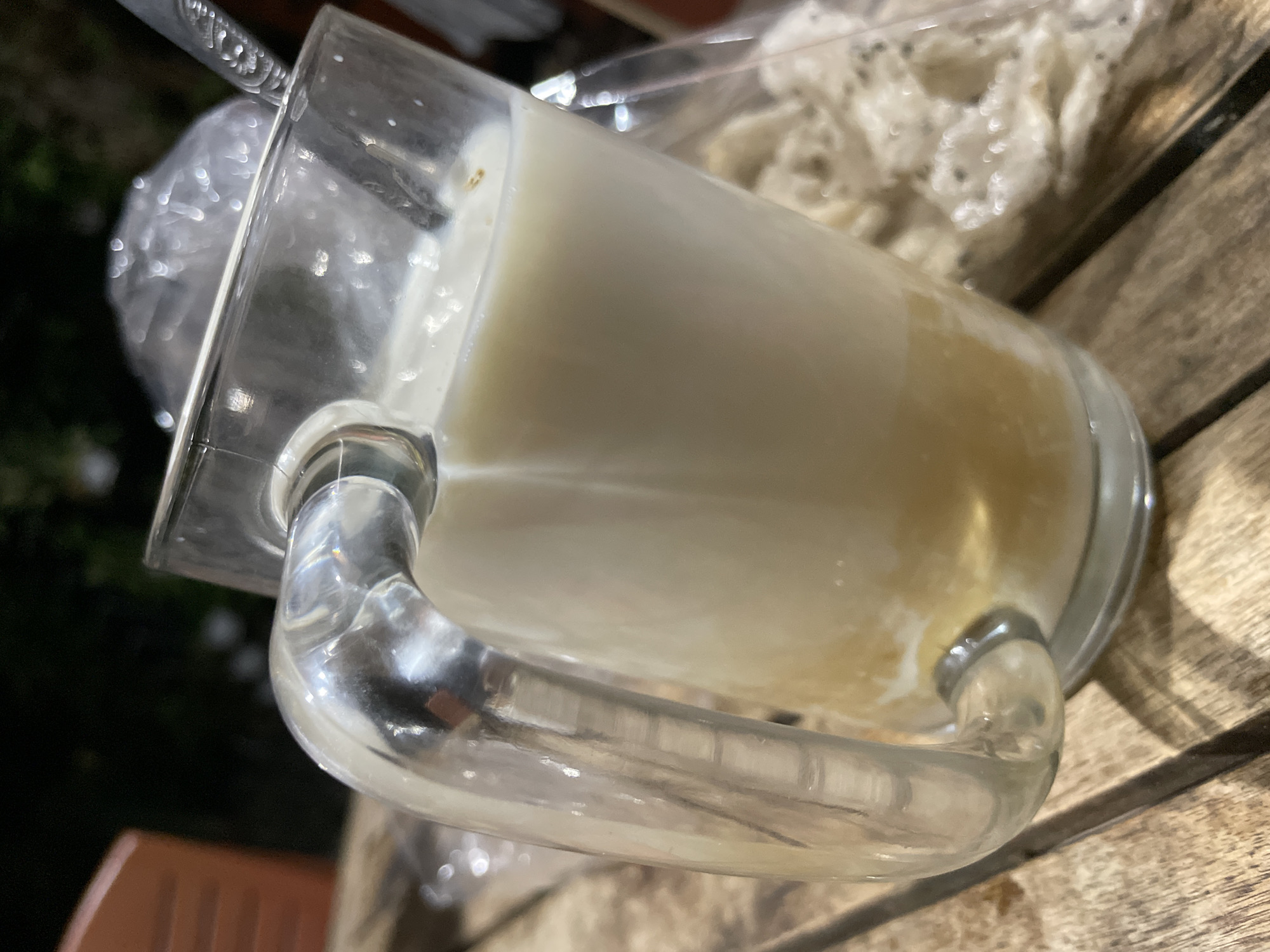A glass mug filled with a milky beverage rests on a wooden table. The mug has a thick, clear glass handle. At the bottom of the mug, a layer of golden liquid is visible beneath the white beverage. A spoon with a patterned handle is submerged in the drink. In the background, a blurred plastic-wrapped item and the dark outline of foliage suggest an outdoor setting, possibly a restaurant or cafe in Phan Thiet Ward, Vietnam. The lighting indicates it may be evening or nighttime.