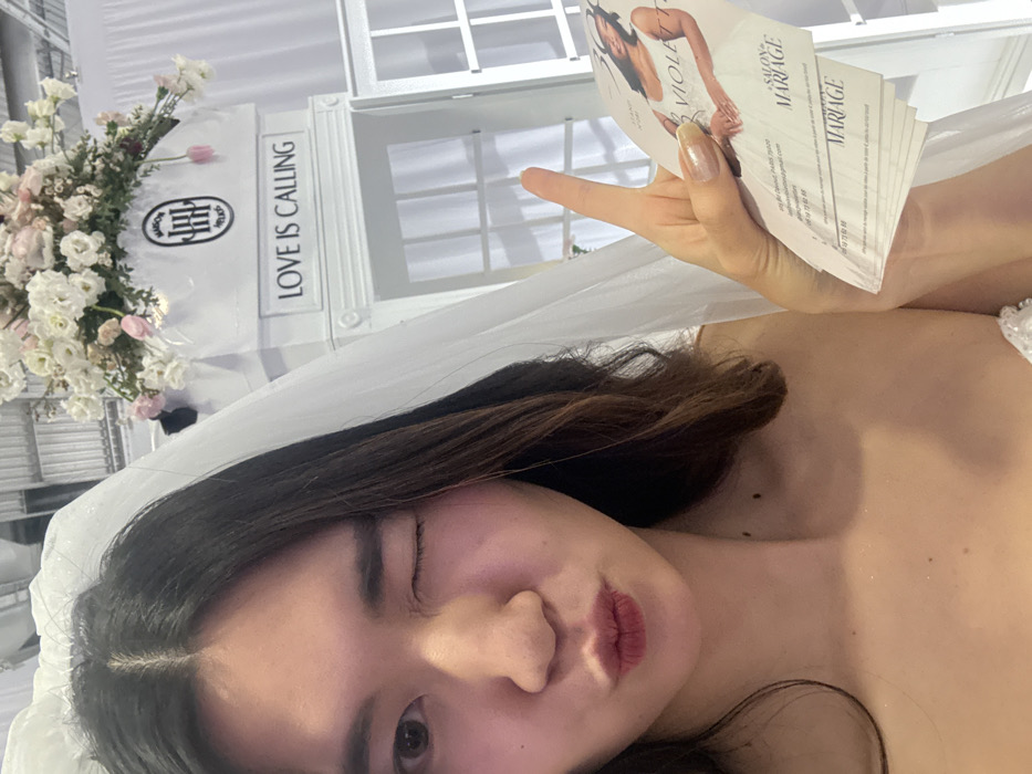 A selfie-style shot captures a woman wearing a white wedding dress and veil. She winks at the camera while holding a stack of business cards from "Salon Mariage" featuring a woman in a wedding dress on the front. A decorative white structure with the phrase "Love is Calling" and floral arrangements sits in the background. The image has a soft, bright, and celebratory feel.