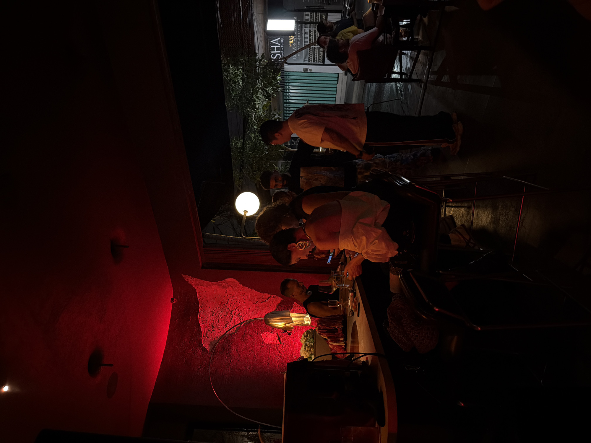 The image depicts a dimly lit indoor scene, possibly a bar or restaurant, with red lighting casting a warm glow on the textured walls. Several people are visible, appearing to be socializing or dining at tables and a counter. In the foreground, a woman with large hoop earrings and a white top is seated, looking at her phone. Next to her, a man in a black tank top is also seated at the counter, seemingly engaged in conversation. Further back, a group of people are gathered around a table, with one man in a light-colored t-shirt leaning forward. Outside, a lit sign for "SHA" is visible, along with greenery and what appears to be a wet street, suggesting it might be nighttime or raining. The overall atmosphere appears casual and relaxed.