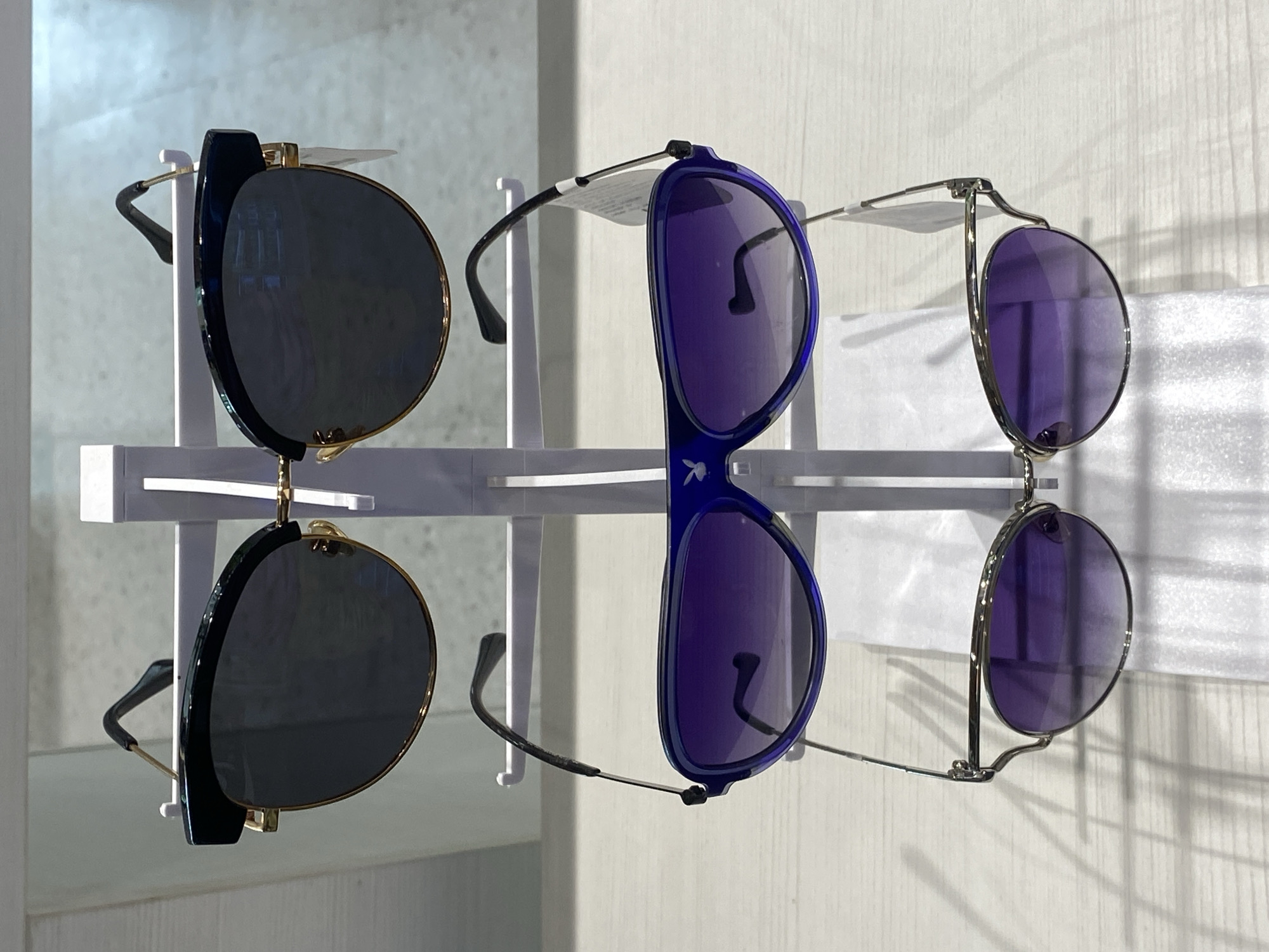 The image shows three pairs of sunglasses displayed on a white rack against a white wall. The sunglasses are of different styles: one pair has black frames and lenses, another has blue frames and purple lenses, and the third has silver frames and purple lenses. The environment appears to be a retail setting. It is unknown if this image was taken in Choeng Thale, Thailand.