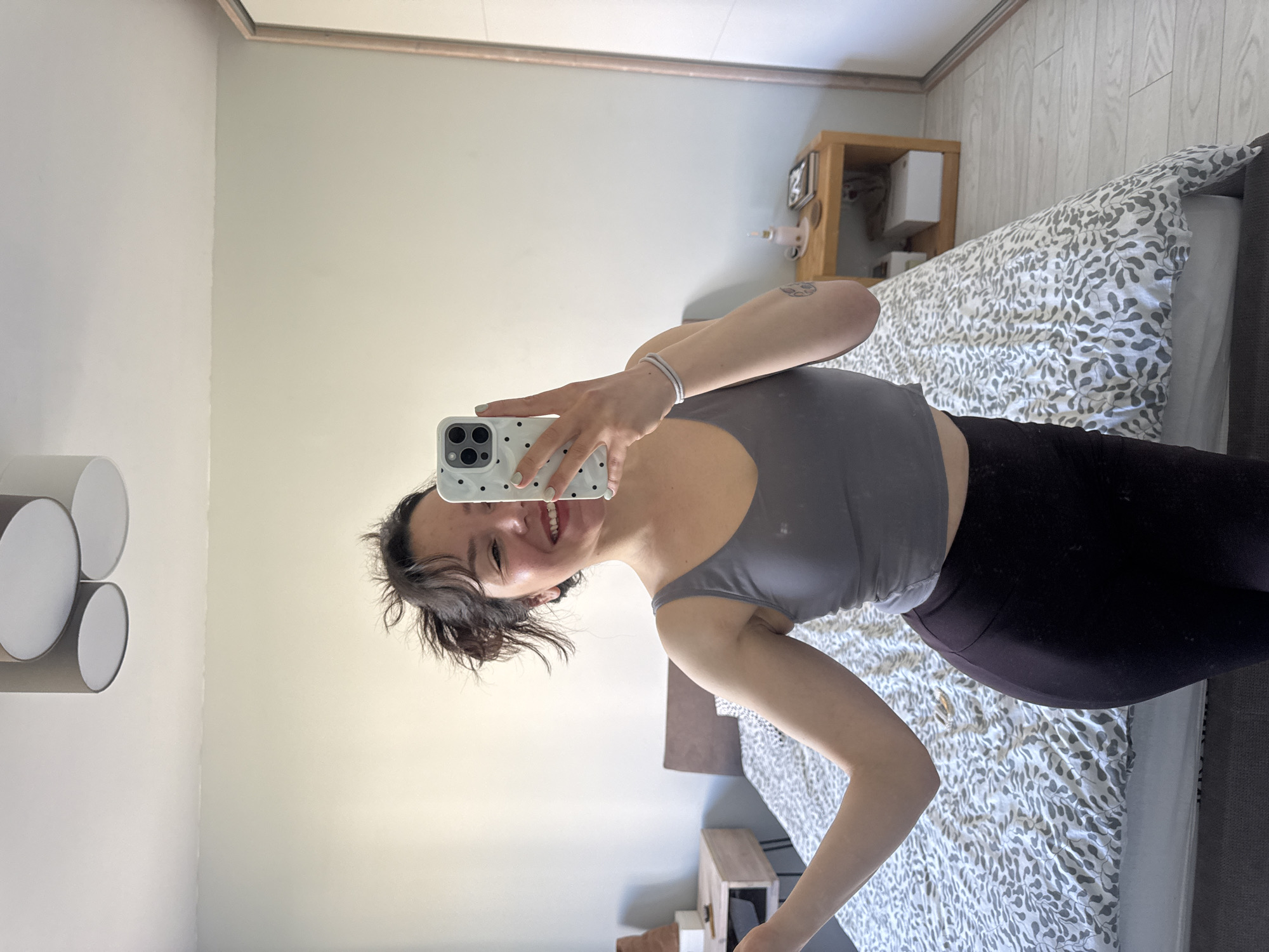 A woman is taking a selfie in a bedroom. She is wearing a gray tank top and black leggings. The bed has a gray and white patterned bedspread. There is a nightstand with items on it next to the bed. There are decorative lights on the wall. The location is likely an interior residential space. The image does not provide any information to definitively place the location in Burgas, Bulgaria.