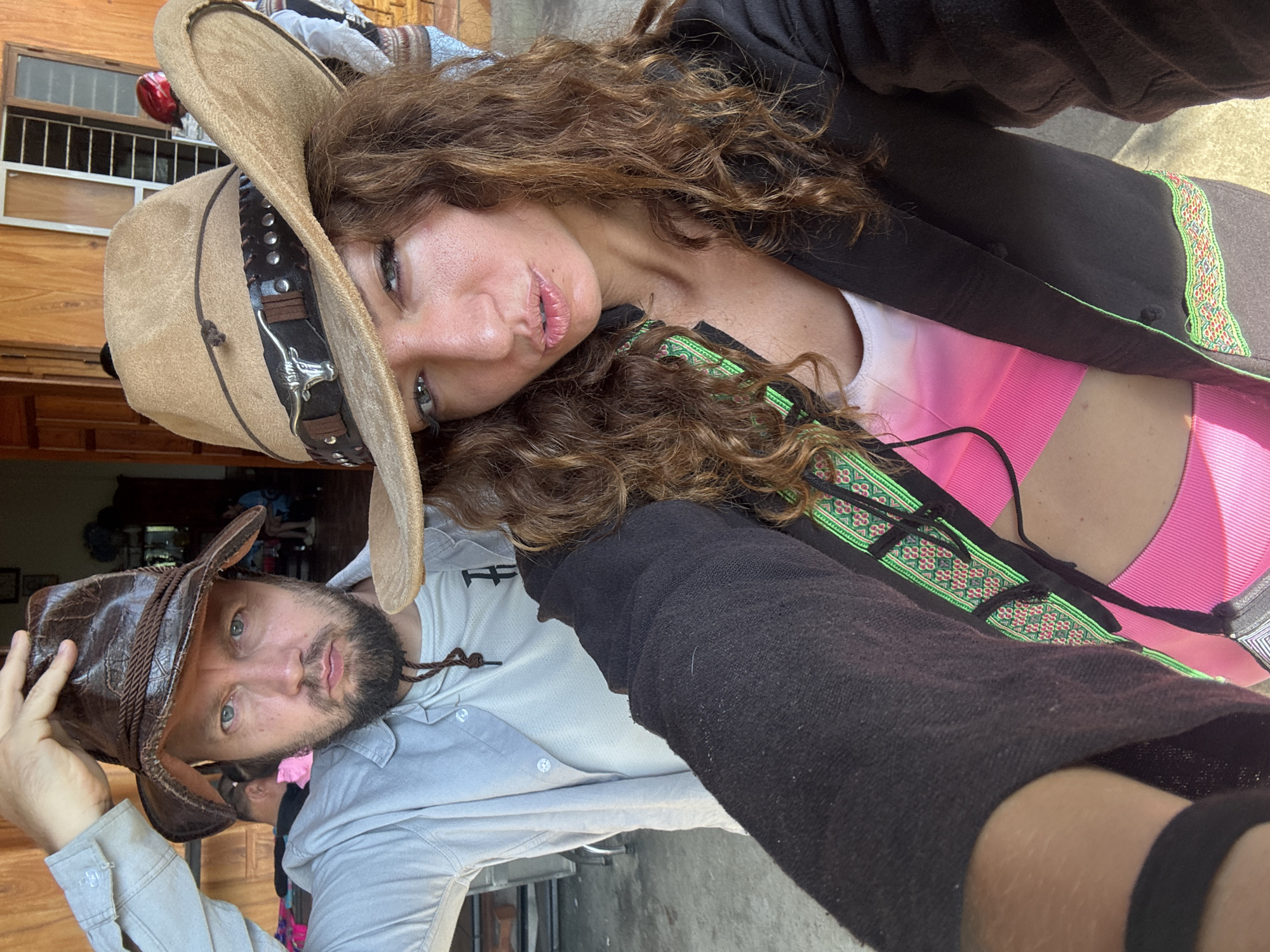 A selfie captures a man and a woman in what appears to be Bắc Hà, Vietnam, judging by the traditional clothing trim visible on the woman's jacket. The woman is wearing a tan cowboy hat with a brown band and a silver steer head adornment, a black jacket with an embroidered green trim, and a pink top. The man wears a weathered brown leather cowboy hat and a light gray button-down shirt. The background shows parts of a wooden building with barred windows and other indistinct objects.