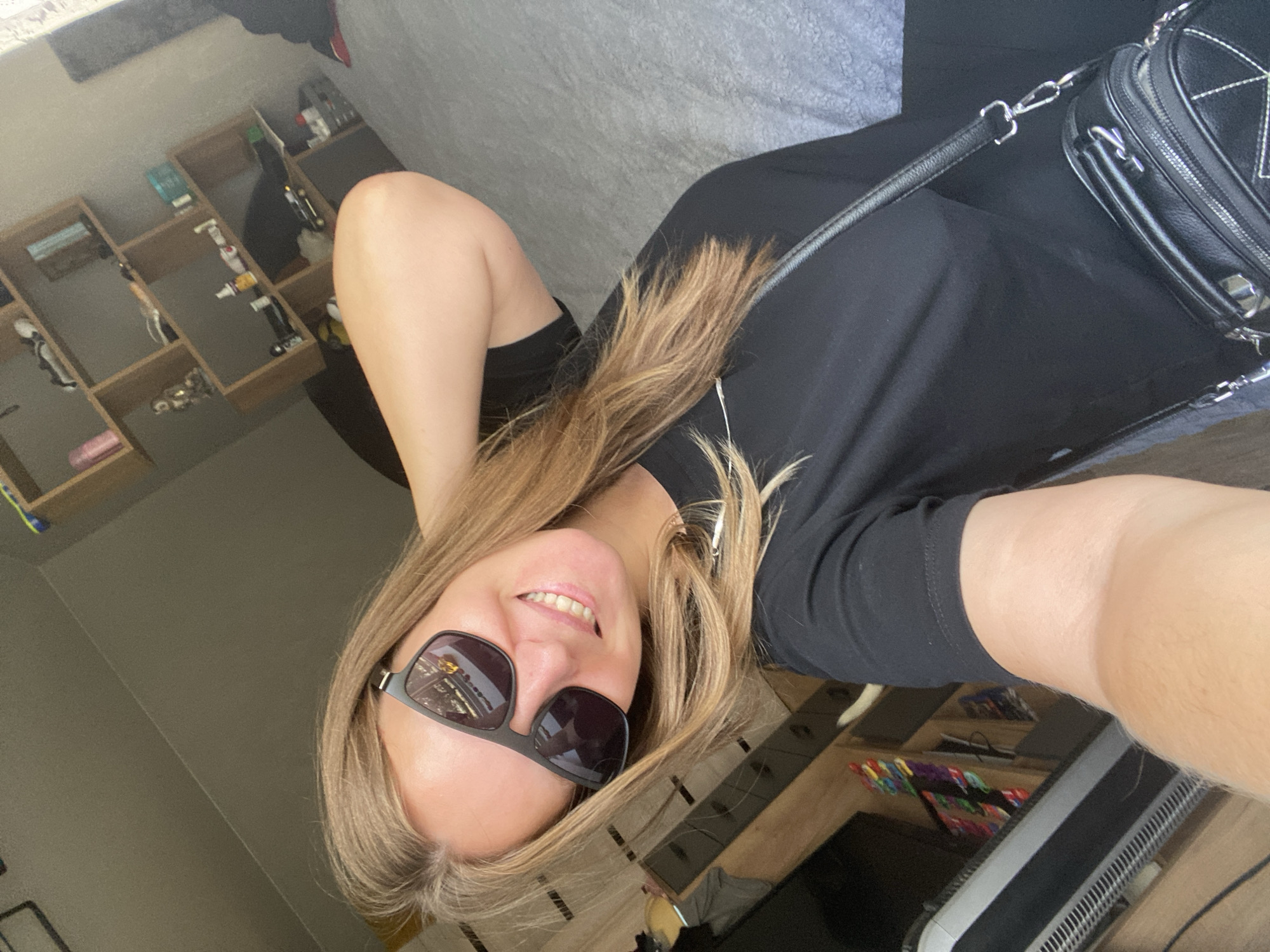 A young Caucasian woman with blonde hair, wearing sunglasses and a black t-shirt, is taking a selfie indoors. She is sitting on a bed with gray bedding, and a shelf unit with various items is visible in the background. A black purse with a strap is slung over her shoulder. The room appears to be a bedroom. There is no information in the image to suggest the location is in Noviy, Russia.