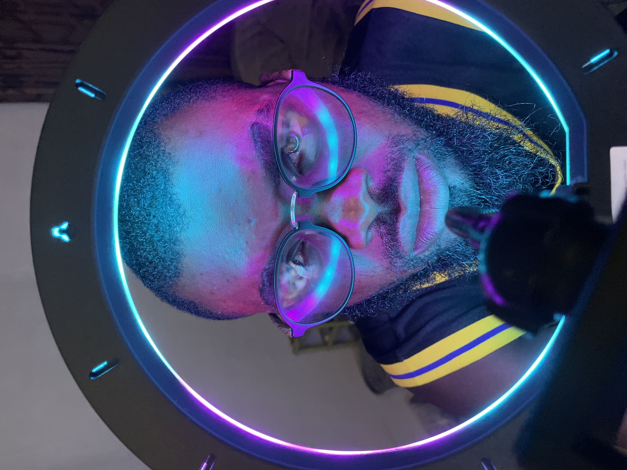 This is a close-up, upside-down shot of a Black man wearing glasses and a black shirt with yellow and purple stripes. He has a beard and is looking into a ring light that is casting vibrant blue and purple light onto his face. The ring light has small, glowing blue indicators on its outer edge. The man appears to be indoors, possibly in a studio or a room with controlled lighting, given the setup of the ring light. The lighting suggests it could be nighttime or a dimly lit indoor environment. The overall mood is artistic and stylized due to the dramatic lighting. No visible text is present in the image.