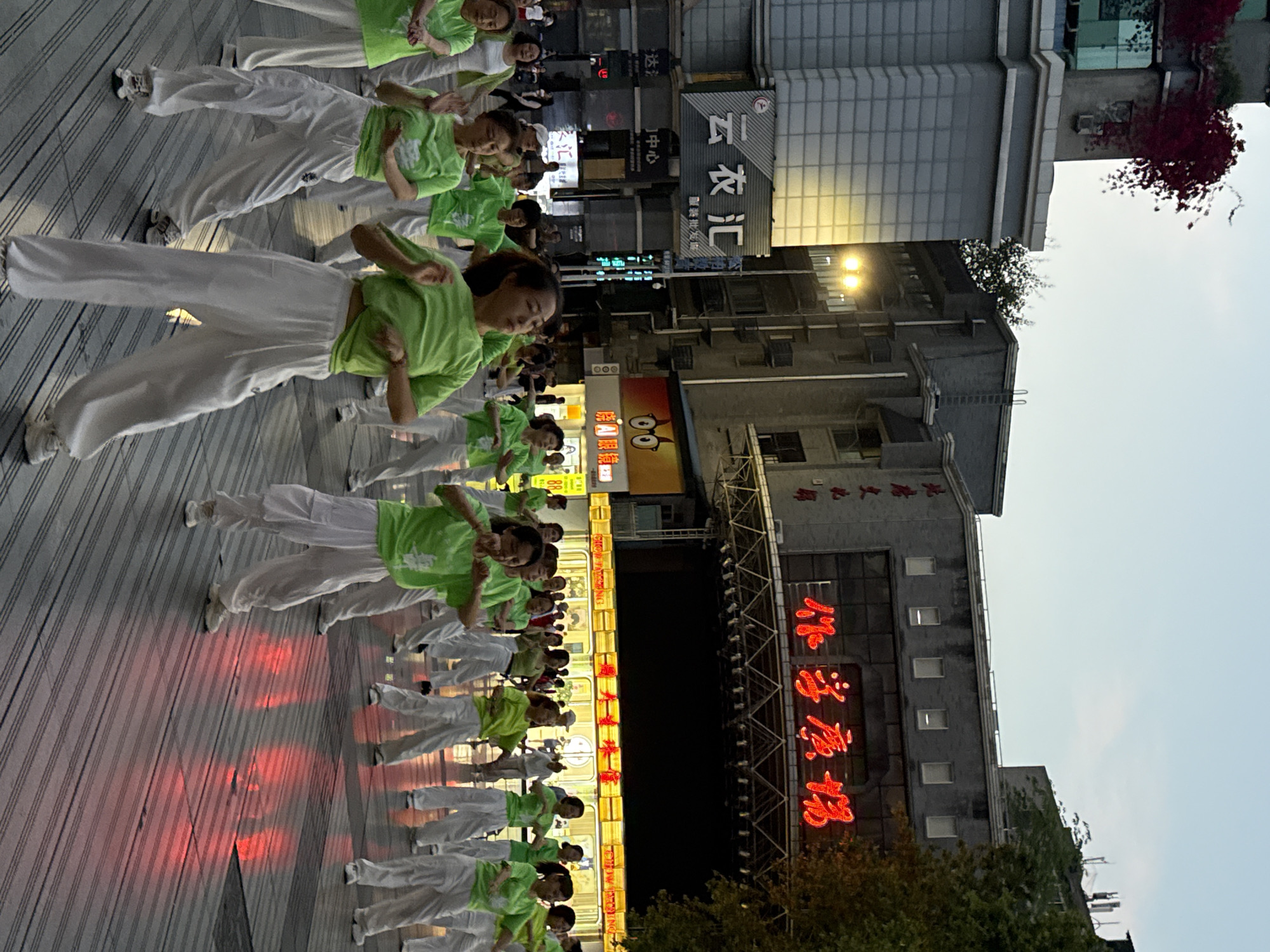 A line of people in white pants and light green shirts are dancing on a wooden platform in Beibei, China, on what appears to be a city street at night. The lighting is dim, but the performers are illuminated by red stage lights and store signs nearby. The street is lined with businesses and tall buildings. A sign for a restaurant and/or business in Chinese is visible in the background.