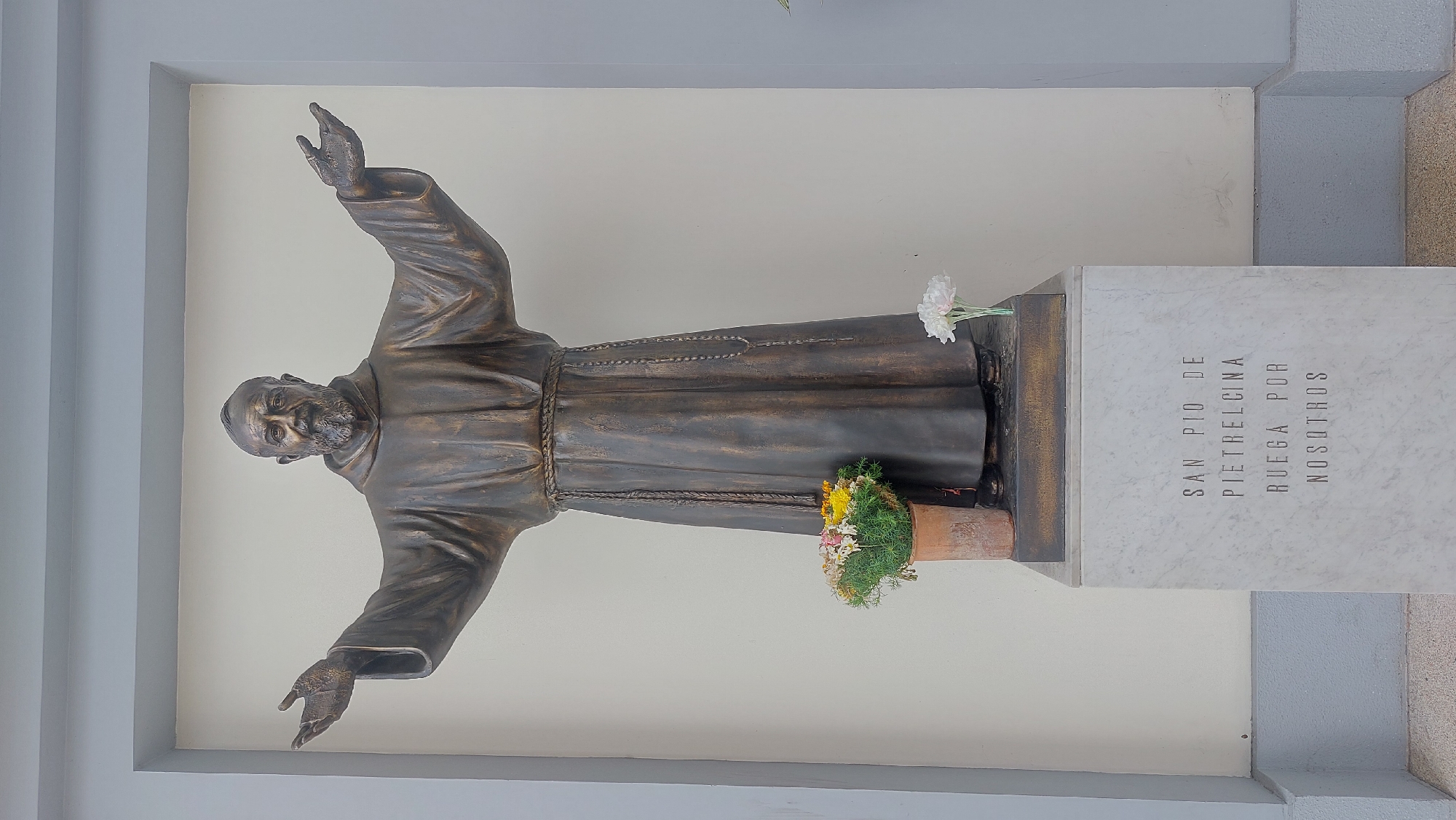 A statue of Padre Pio de Pietrelcina stands within what appears to be a niche or recessed area, possibly a devotional alcove. The statue is bronze-colored, depicting a man in a robe with his arms outstretched. A small pot with flowers sits at the base of the statue on the left side. The statue rests on a stone pedestal, on which is engraved the text "SAN PIO DE PIETRELCINA RUEGA POR NOSOTROS". The statue is set against a plain off-white background, framed by architectural elements, possibly part of a building facade. The location is the city of Caracas, Venezuela.