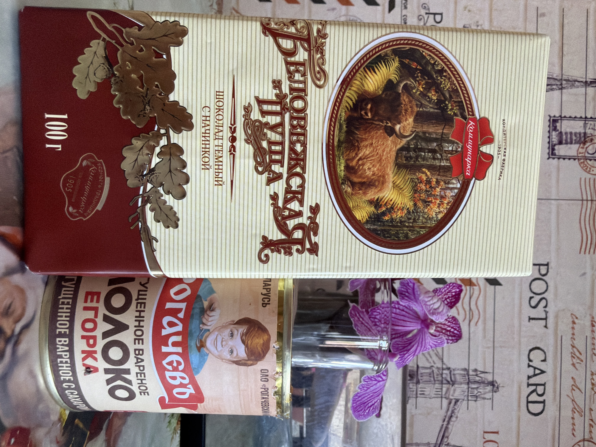 The image displays a tabletop scene with various food items and a floral arrangement. There is a rectangular package of "Belovezhskaya Pushcha" dark chocolate with filling from the confectionery factory "Kommunarka" (established in 1905). Next to it, is a can of "Rogachev" boiled condensed milk (with the name "Egorka" on it). A vase holding purple orchids is positioned between the can and the chocolate package. The entire scene is set against a backdrop printed with vintage postcard imagery. The location is not directly visible. The city of Chaikovsky, Russia, is not mentioned.