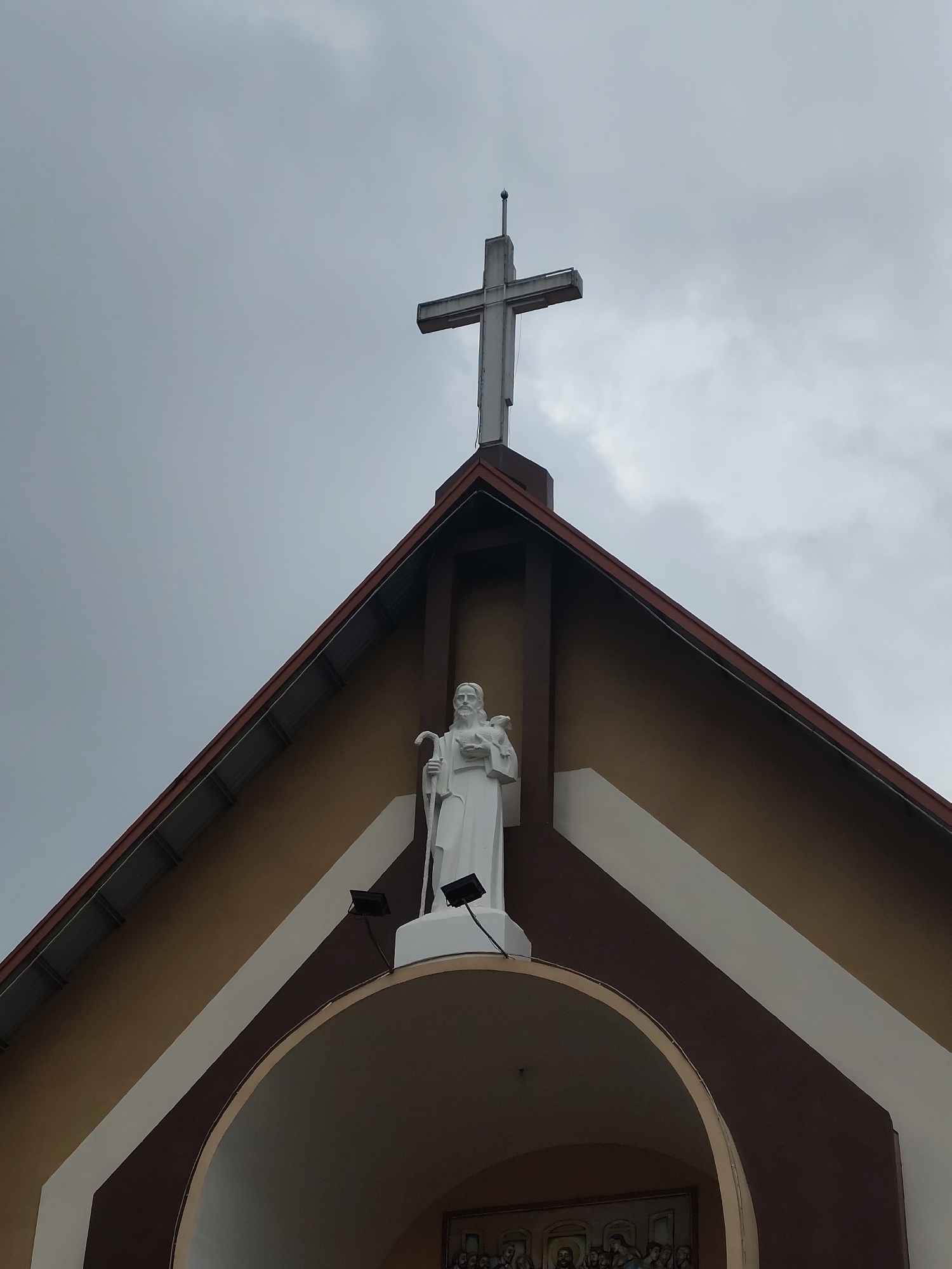 This upward-angled shot captures the upper facade of a church or religious building, located in Lang Biang - Đà Lạt, Vietnam, under an overcast sky during daytime. The scene is static, focusing on architectural and religious elements.
Dominating the pinnacle of the gable roof is a large, weathered concrete cross, stark against the grey, cloudy sky. Directly below the cross, positioned within a recessed section of the gable, stands a white statue of a bearded male figure. This figure, likely a saint or Jesus as the Good Shepherd, holds a staff in one hand and a lamb resting on his other arm. Two black spotlights are mounted on either side of the statue, presumably for nighttime illumination.
The building's gable features a distinct color scheme: a warm beige for the main wall, bordered by darker brown structural elements and a prominent white diagonal band that runs along the sloped roofline. The roof itself has visible corrugated eaves. Below the statue, an arched entryway or niche is partially visible. Inside this archway, a painting or mural can be faintly seen, depicting multiple figures seated at a table, strongly suggesting a representation of "The Last Supper" or a similar biblical scene. The overall impression is one of solemnity and religious significance, typical of a Christian place of worship.