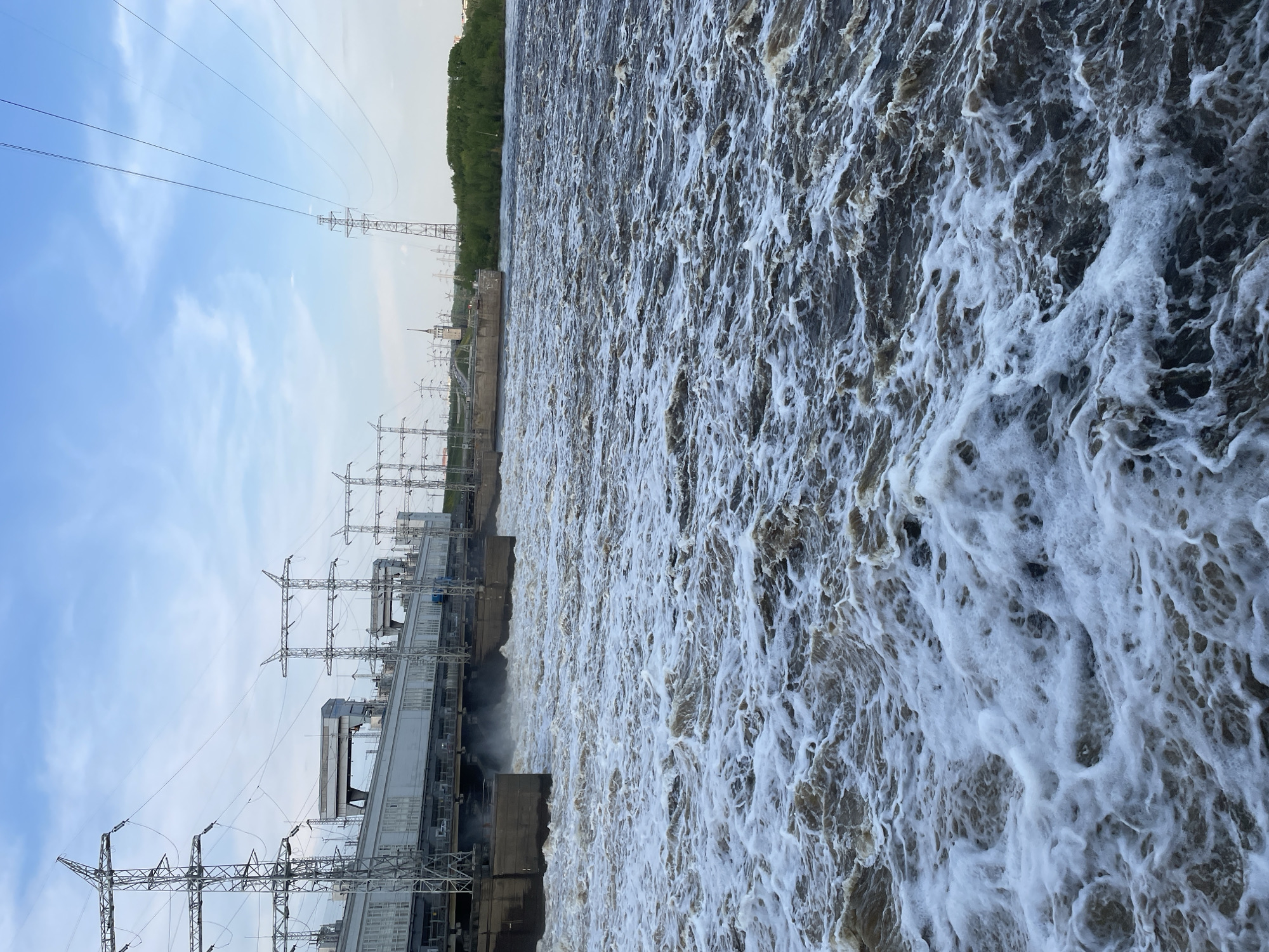 The image shows the Kama Hydroelectric Station in Perm, Russia. Turbulent water is flowing rapidly and vertically downwards. A power station with pylons and power lines is visible on the left. There is a line of trees on the horizon, and a blue sky with some clouds is visible at the top left.