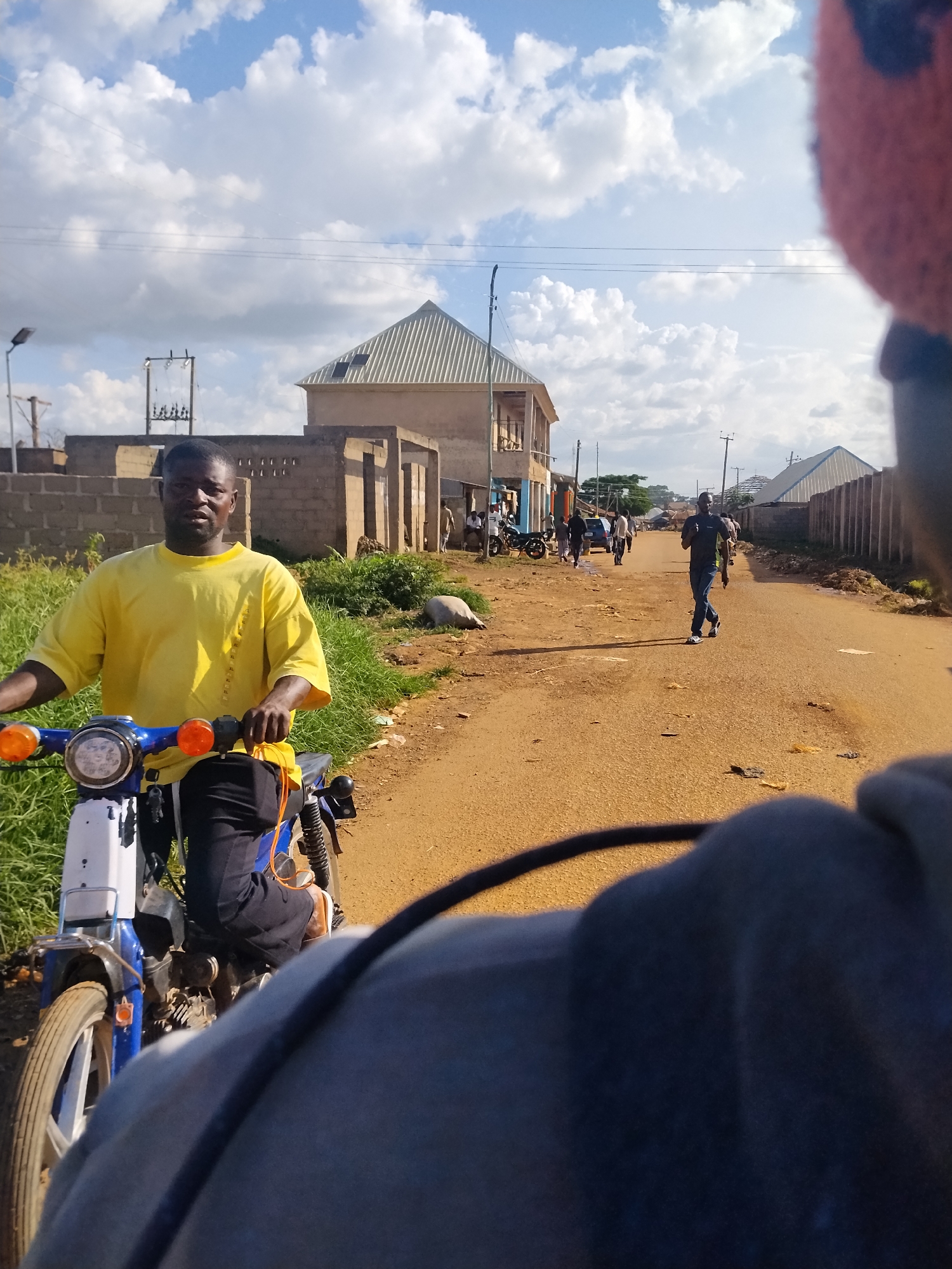 The image depicts a street scene in Mangu Town, Nigeria, under a cloudy sky. In the foreground, a person wearing a blue and white motorcycle jacket is partially visible from the shoulders up, looking out at the scene. The motorcycle itself is blue and white, with orange turn signals. The driver of the motorcycle, a man wearing a yellow t-shirt and black pants, is looking directly at the camera. He is positioned on the left side of the frame.
The street is unpaved and appears to be made of dirt and gravel. On either side of the street are various buildings, some appearing to be under construction with exposed bricks and concrete. There are also houses with corrugated metal roofs. In the mid-ground, several people are walking along the street and standing in front of buildings. One man is walking away from the camera towards the right side of the frame. Further down the street, more people are visible, some gathered near motorcycles and cars. The environment appears to be a developing area within the town, with a mix of residential and commercial structures. Power lines are visible running overhead. The overall impression is one of a typical day in a Nigerian town.