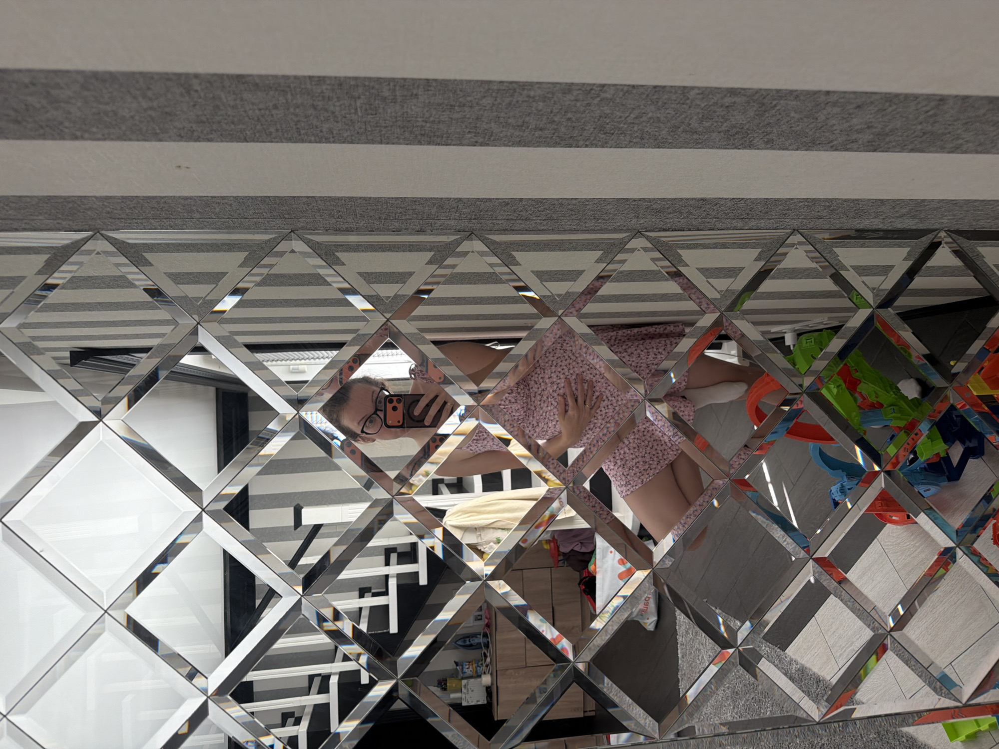 A woman is taking a mirror selfie. She is wearing glasses and a pink dress with a floral pattern. She is holding her belly with her right hand. The mirror is made of faceted diamond-shaped pieces and reflects a room with striped wallpaper. A staircase is visible in the background. The floor is tiled. There are toys on the floor. The image is taken from a low angle.