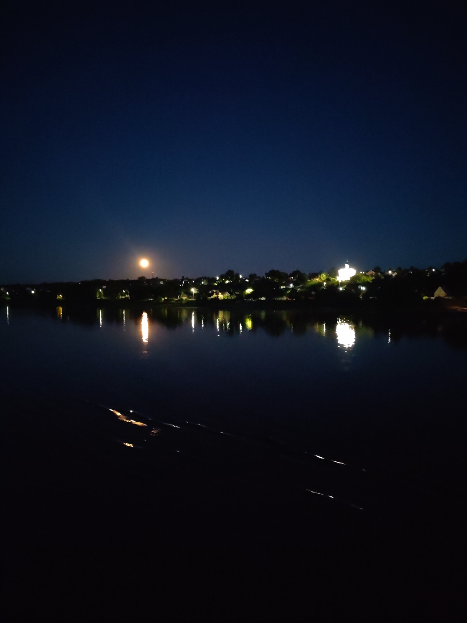 The image depicts a night view of the Volga River in Tutaev, Russia. A large, bright full moon is visible in the dark blue sky, casting a soft glow. Along the riverbank, a line of trees and buildings with illuminated windows is silhouetted against the night sky. The lights from the town are reflected in the dark, still water of the river, creating shimmering patterns. The foreground of the image is dark, with some subtle reflections on the water's surface, possibly from lights or movement within the water.