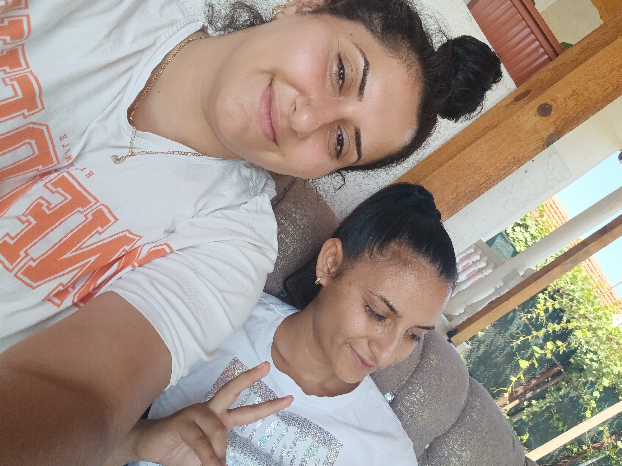 The image shows two women, likely in their late 20s or early 30s, posing for a selfie outdoors. The woman in the foreground, on the left, has dark, curly hair pulled up in a bun and is looking directly at the camera with a smile. She is wearing a white t-shirt with orange lettering and a gold necklace. The woman in the background, on the right, has dark hair pulled back in a ponytail and is looking off to the side with a slight smile. She is wearing a white t-shirt with a sequined design. They are both seated on a cushioned outdoor sofa. The environment suggests a residential area, with visible wooden beams, a white building, and lush green foliage in the background. The presence of the wooden beams and the style of the building's exterior, along with the surrounding greenery, are consistent with elements found in the village of Zimnitsa, Bulgaria.