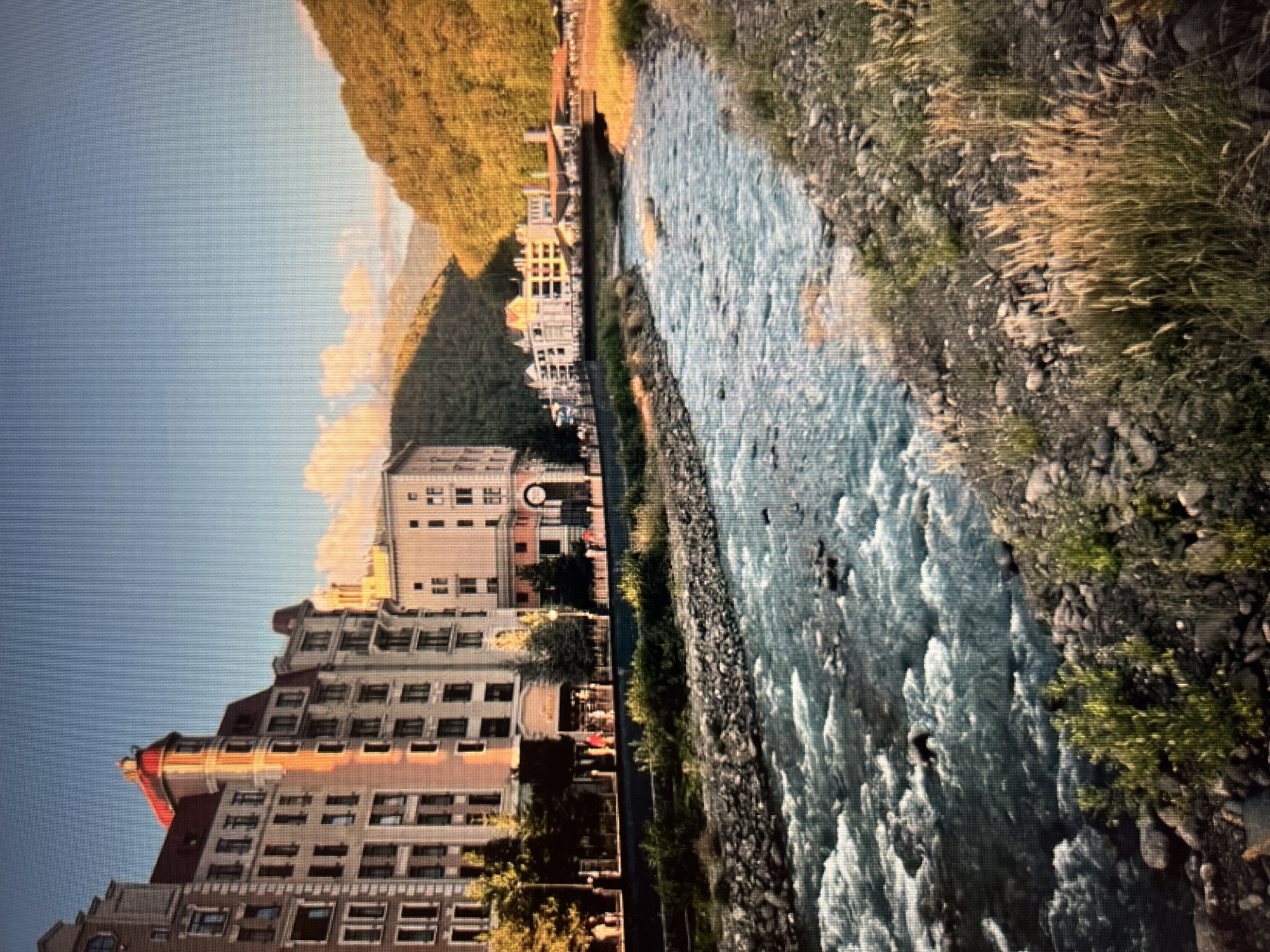 The image depicts a scenic view of a river flowing through a town nestled in a valley. In the foreground, a rocky riverbed is visible, with the water appearing turbulent and clear. The riverbanks are lined with stones and some sparse vegetation, including tall grasses.
To the left of the river, a row of multi-story buildings with light-colored facades and red-tiled roofs are situated along a promenade. People can be seen walking along this promenade. These buildings have a distinct architectural style, with numerous windows and decorative elements.
In the background, a lush green mountainside dominates the scene, with trees covering its slopes. The sun appears to be setting or rising, casting a warm glow on the mountains and clouds. A bridge crosses the river further in the distance, connecting to more buildings on the other side.
While the prompt asks for the location in the city of Khimki, Russia, the visual elements in the image do not specifically confirm this location. The architecture and natural landscape could be found in various mountainous regions.