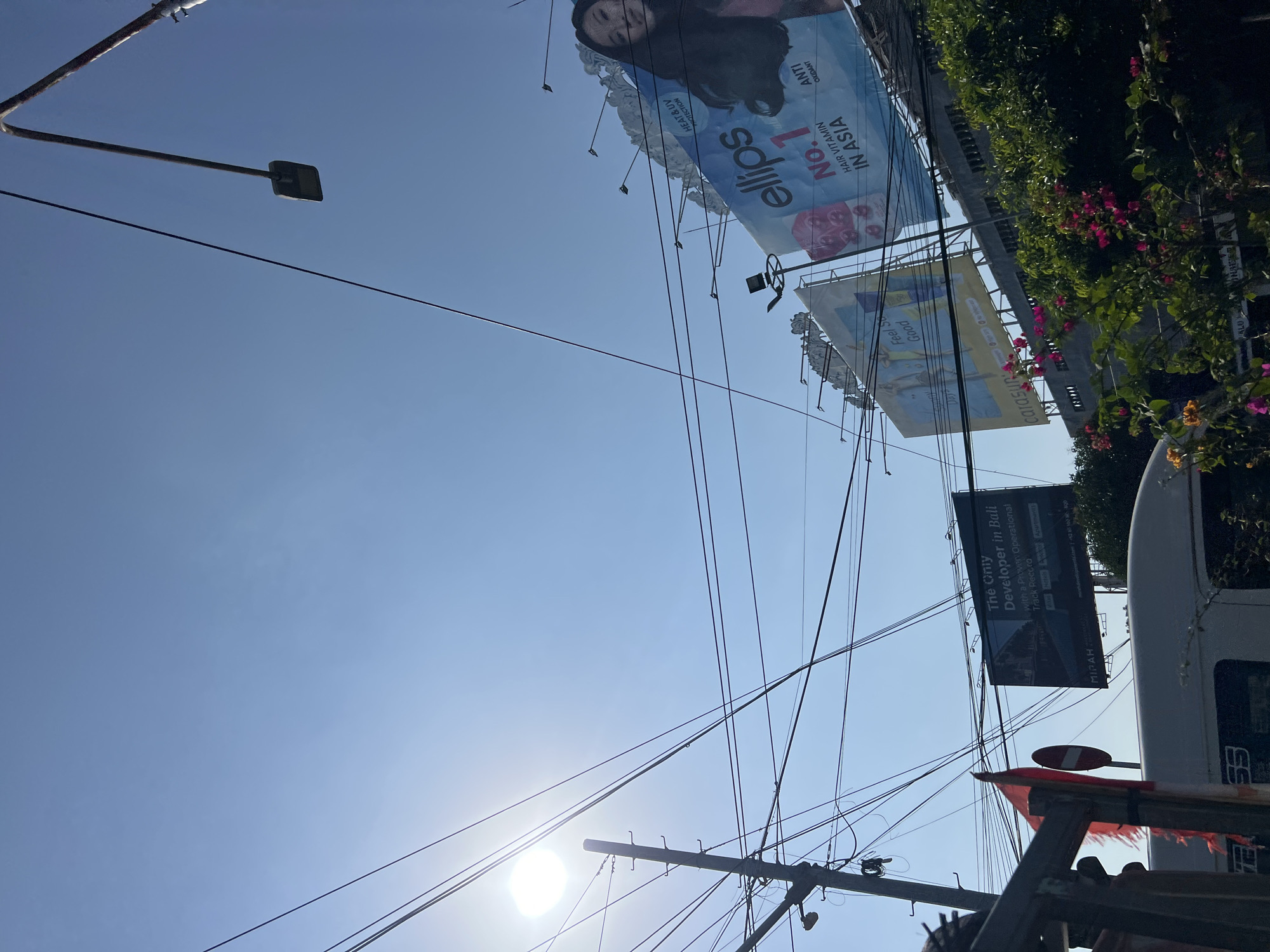 The image depicts a street scene in Denpasar Barat, Indonesia, on a clear, sunny day. The foreground is dominated by a dense network of overhead electrical wires, strung between utility poles. The sun, high in the sky, creates lens flare and brightens the scene.
Several billboards are visible. The largest, on the right, advertises "ellips No. 1 HAIR VITAMIN IN ASIA" with an image of a woman. To the left of this, another billboard promotes "carasun," featuring stylized figures and what appears to be sunscreen. A third billboard, further to the left, reads "The Only Developer in Bali With a Proven Operational Track Record," displaying an image of a modern building.
A utility pole with multiple crossbars and insulators is prominently positioned on the left, with its wires converging above. A street lamp on a curved arm extends from the upper right of the frame.
In the lower left, a red and white Indonesian flag is partially visible, mounted on a dark structure. A white vehicle, likely a bus or van, is present at the bottom, with some of its markings obscured. Lush green foliage and bright pink bougainvillea flowers are visible along the bottom edge of the image, partially obscuring the lower parts of the billboards and the vehicle. The overall impression is a busy urban streetscape with commercial advertising set against a backdrop of utility infrastructure and natural elements.