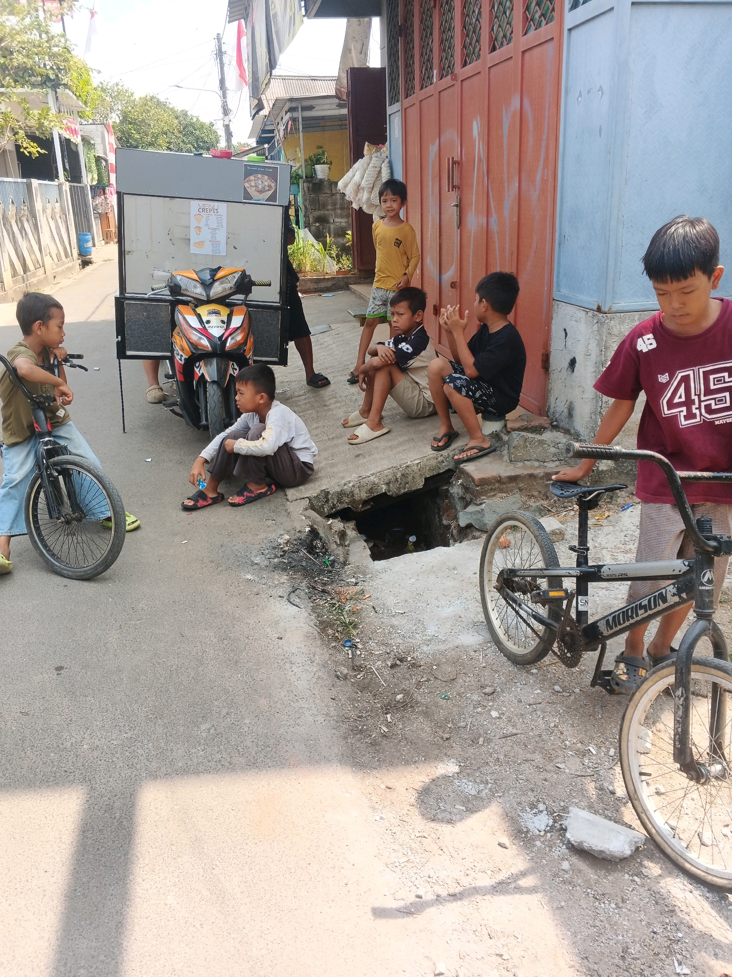 In the city of Kadu, Indonesia, a street scene unfolds under a clear, sunny sky. In the foreground, a young boy on a bicycle, wearing blue jeans and a khaki shirt, looks towards the left. Next to him, another boy sits on the ground, dressed in dark pants and a light long-sleeved shirt, holding a blue object. Behind them, a motorcycle is attached to a food cart. To the right, a group of boys are gathered. One boy in a yellow shirt stands, while two others are seated. The eldest boy, wearing a maroon shirt with the number "45" on it, stands beside a bicycle, looking down. The environment is a paved street with a damaged section near a drain. Buildings with orange and blue walls form the backdrop.