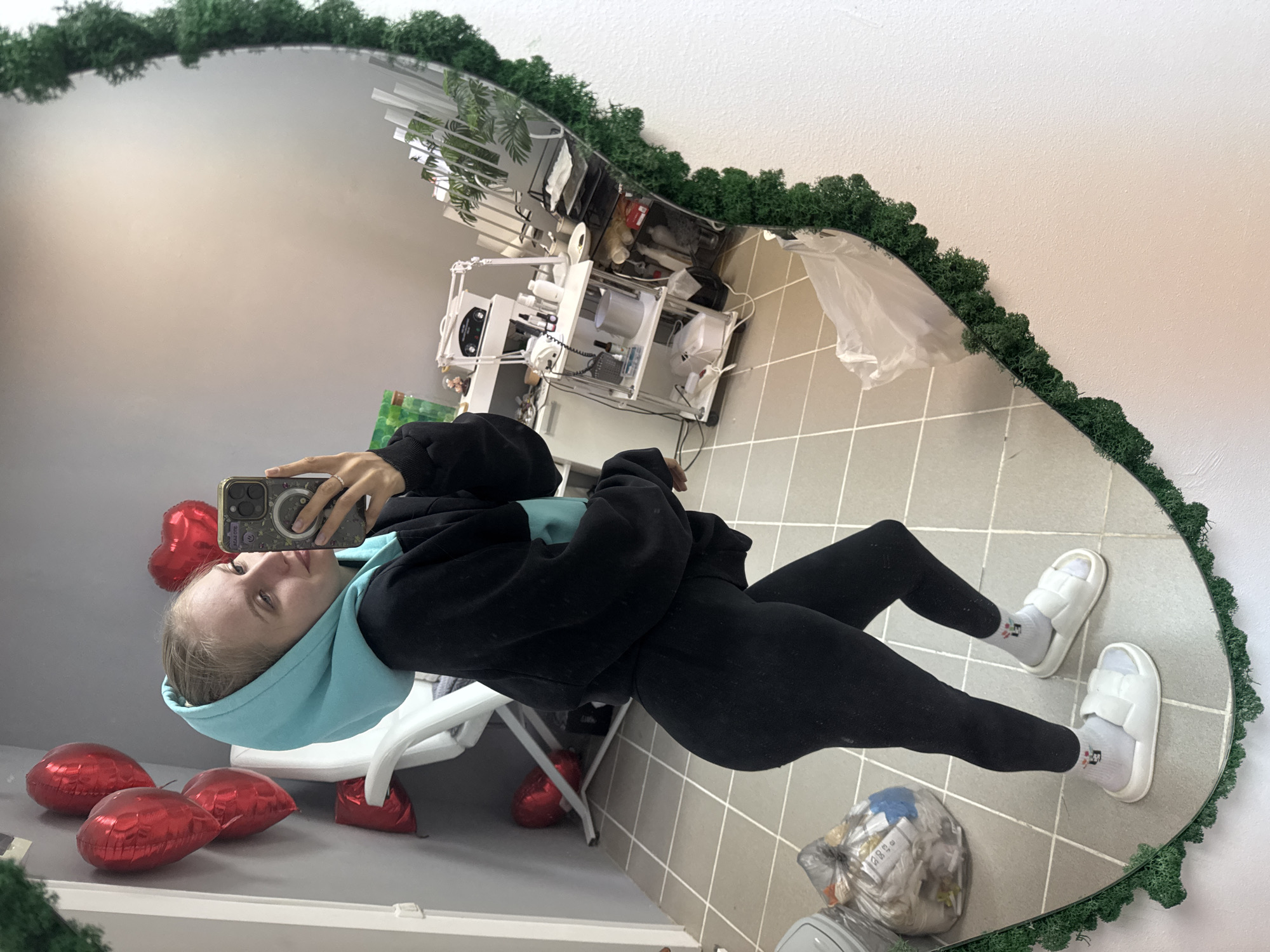 A young woman is captured taking a mirror selfie in what appears to be an indoor commercial space in Izhevsk, Russia. The image is a reflection in a large, heart-shaped mirror framed with green, moss-like material.
The woman is dressed in a black hoodie with a light blue lining in the hood, black leggings, white socks featuring a small black and red cartoon-like pattern, and white slide sandals. She holds a dark-cased smartphone, possibly an iPhone, in her right hand, positioned to capture her reflection. Her face is visible in the mirror, showing light hair and a neutral expression as she looks towards the phone screen.
On the tiled floor to her left, several deflated or partially inflated red, heart-shaped foil balloons are scattered. A white chair with a metallic frame is visible behind her legs. To her right, a clear plastic trash bag filled with white refuse is situated on the floor, with some small, illegible text visible on wrappers inside, such as "5300" and partial Cyrillic characters like "КЭ" or "КУ", along with "115/150".
The background reflected in the upper part of the mirror reveals white walls, a light-colored ceiling, and various pieces of professional-looking white and metallic equipment, suggesting the setting could be a beauty salon, a medical office, or a specialized studio. The presence of heart-shaped balloons hints at a celebratory or themed decoration. The lighting is bright and even, indicating an indoor environment, likely during the daytime or in a well-lit interior.