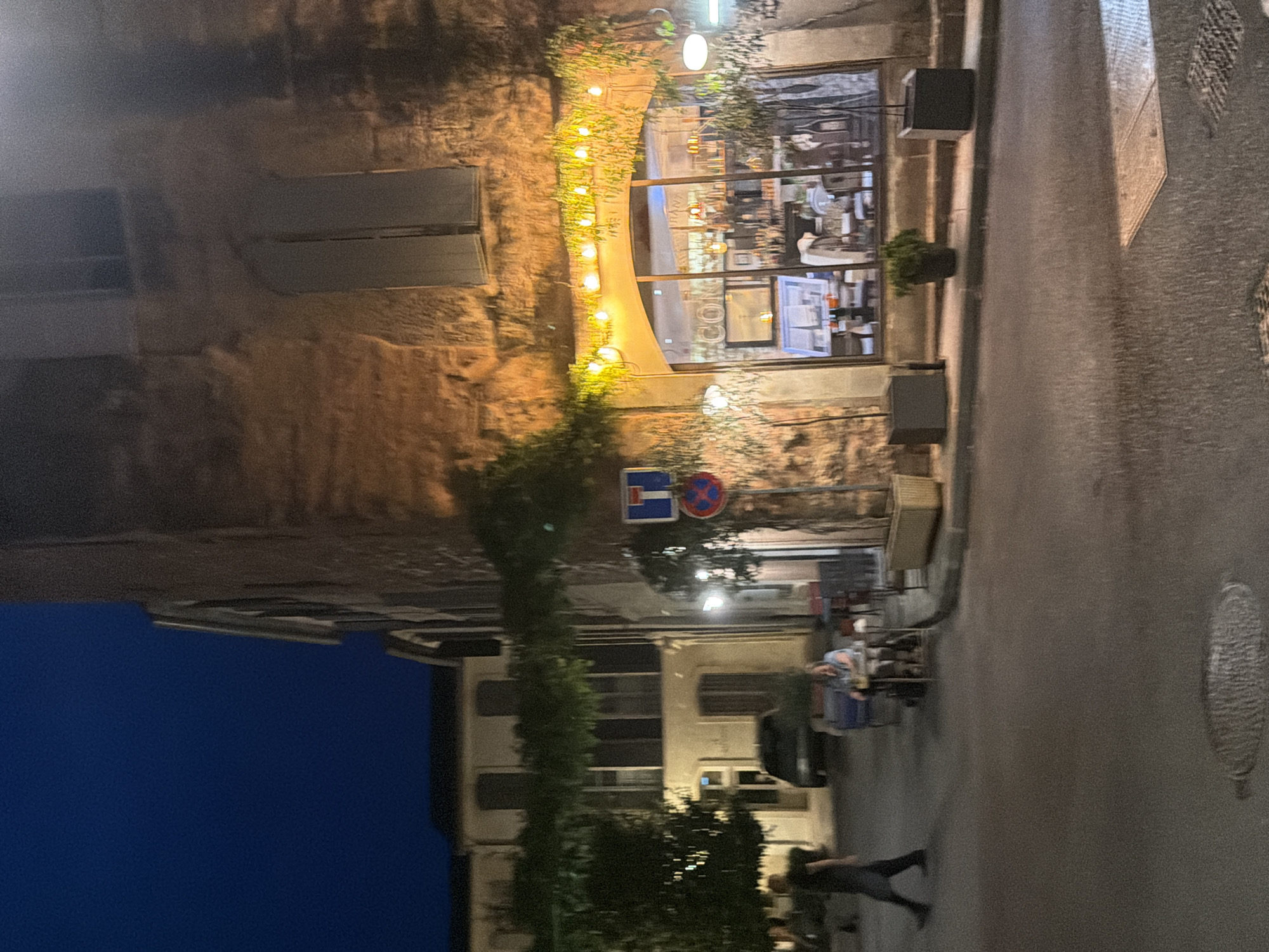 The image depicts a street scene in the city of Arles, France, at dusk or night. A building with a rough stone facade dominates the right side of the frame, featuring large arched windows that reveal a brightly lit interior of what appears to be a restaurant or cafe. String lights are draped across the top of the archway, and greenery wraps around the building.
In the foreground, the street surface is visible, with a manhole cover present. To the left of the building, another, lighter colored building is visible, with a tree and some greenery in between. A person is walking away from the viewer on the left side of the street. Further down the street, a person is seated at a small table outside a building, possibly a cafe, with a drink in front of them. There are also some chairs and tables set up on the sidewalk. Two traffic signs are attached to a pole on the right side of the street. The sky is a deep blue, indicating nighttime.