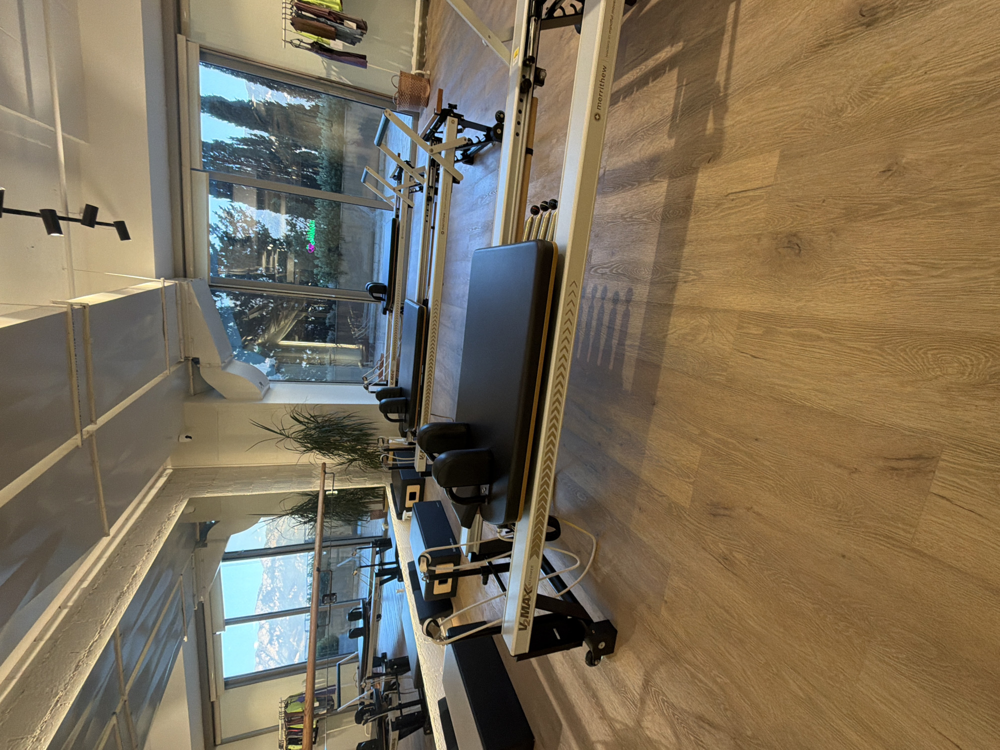 The image depicts the interior of a well-lit pilates studio with large windows offering a view of mountainous terrain and trees. Several pilates reformers are arranged on a wooden floor. In the foreground, a Merrithew V-MAX Reformer is prominently featured, with its black padded carriage, springs, and metal frame visible. Other reformers are seen in the background, some with black padded headrests and handles.
The studio features a reflective wall on the left, likely a mirror, which shows more of the studio's equipment and a barre along the wall. Plants are placed in pots near the windows, and there are racks of clothing visible on the right side of the image. The ceiling has exposed beams and lighting fixtures. The overall impression is one of a modern and spacious fitness facility.
This pilates studio is located in Boreti, Montenegro.