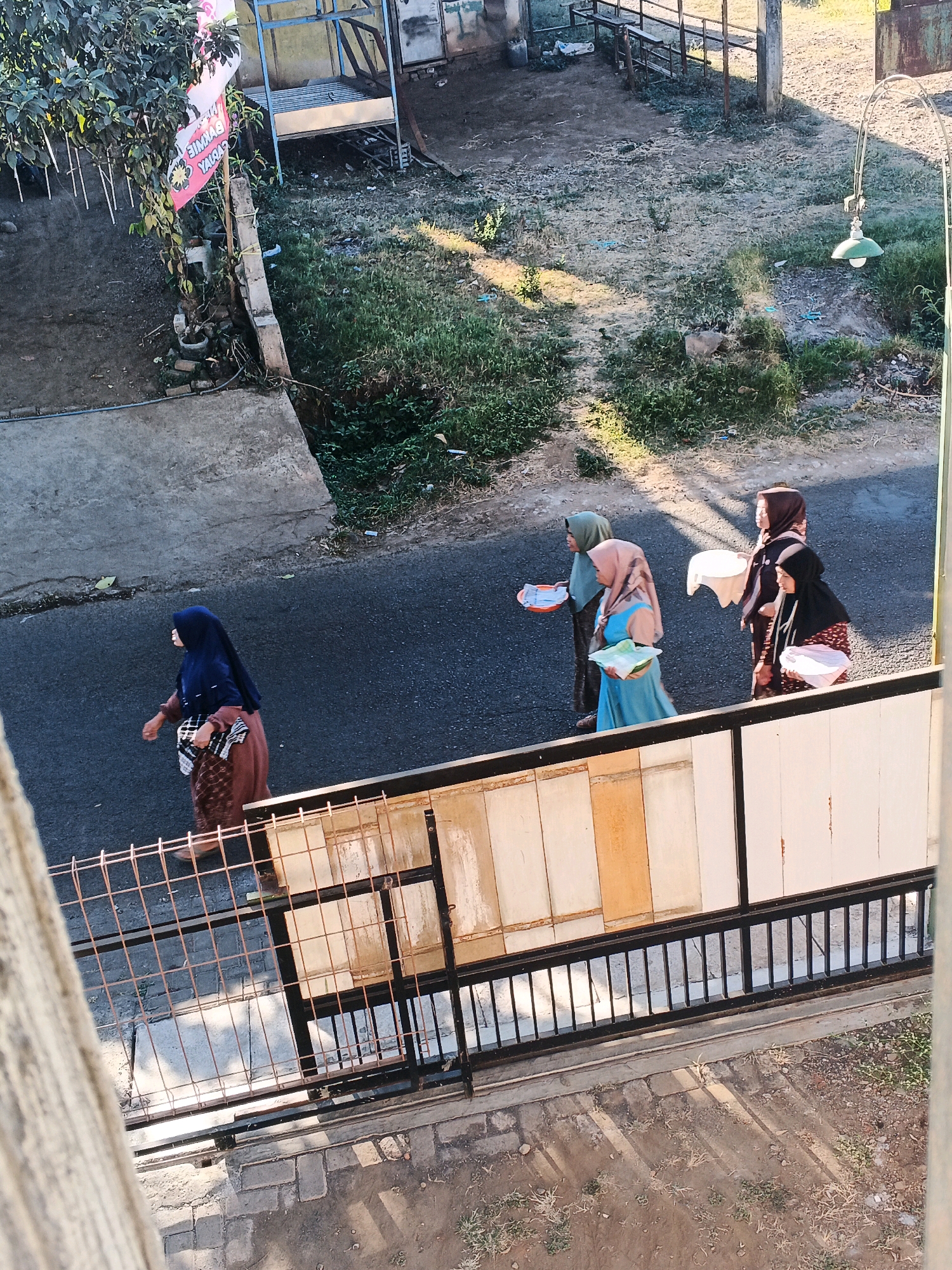 In Bocek, Indonesia, four women walk along a paved road. The woman on the far left wears a dark blue hijab and a brown dress, carrying a checkered bag. To her right, a woman in a light pink hijab and a blue dress carries a green item. Next, a woman in a brown hijab and a dark top holds a white object. The woman on the far right wears a black hijab and a patterned dress, also carrying a white item. They are walking in the same direction, towards the right of the frame. The environment appears to be an outdoor setting with grassy areas and some debris visible. In the background, there is a building with metal structures and a green lamp hanging from a pole. A fence runs along the lower portion of the image.