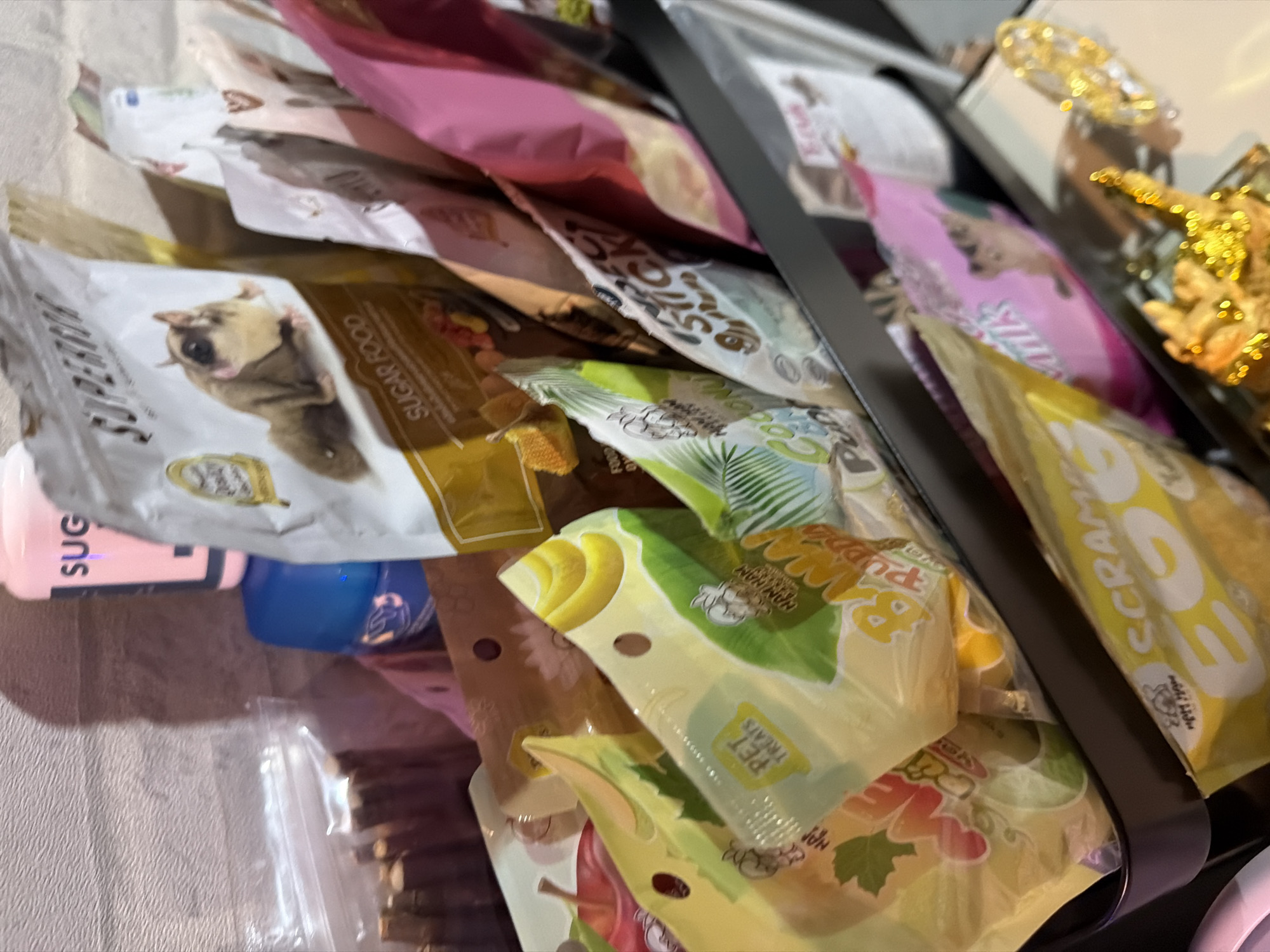 The image shows a collection of pet food bags and treats arranged in a dark colored rack. The bags are of various sizes, colors, and designs, with some featuring images of animals and fruits. Some of the visible text on the bags includes "SUGAR FOOD," "PET TREATS," "BANANA PUPPIES," "STICK," and "SCRAMBLE EGG." In the background, there are some plastic containers and a textured wall, possibly brick. The lighting in the scene is artificial and casts shadows. The overall impression is of a retail display or a personal collection of pet supplies. There is no information in the image to indicate its location in Bangkok, Thailand.