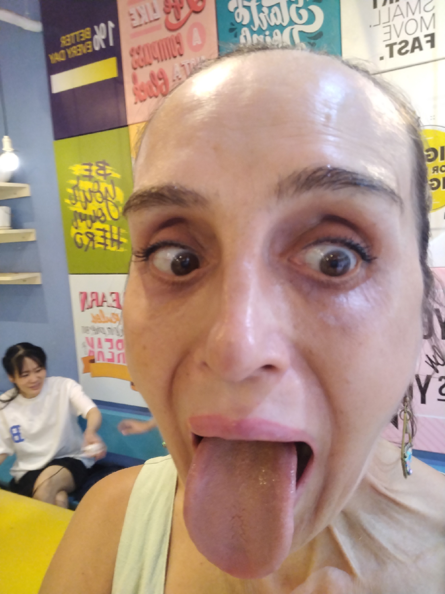 A close-up selfie shows a middle-aged woman with her tongue extended, eyes looking cross-eyed. She is wearing a light-colored top and an earring is visible on the right side of the frame. In the background, a young Asian woman is seated at a yellow table, wearing a white T-shirt with a blue "B" on it and black shorts. The background also includes colorful wall decorations with text and designs. Based on the location tag, this scene is set in Bai Chay Ward, Vietnam.