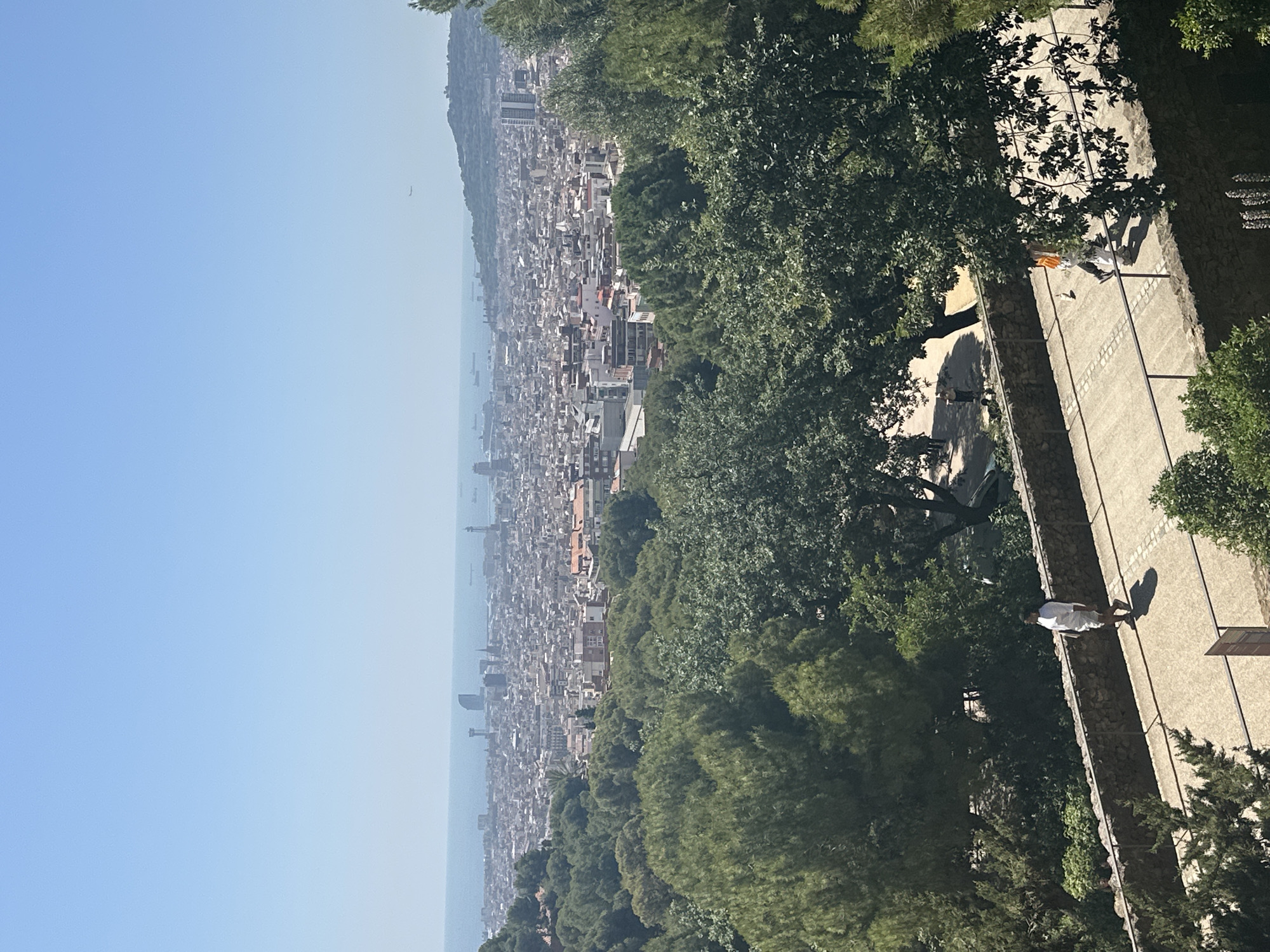 The image captures a panoramic view of Barcelona, Spain, seen from a high vantage point. In the foreground, a pathway lined with a stone wall curves through lush green trees. A person dressed in white walks along the path, casting a shadow on the ground. Further along the path, another individual wearing an orange backpack is visible. Beyond the trees and pathway, the cityscape of Barcelona unfolds, a dense collection of buildings stretching towards the sea. The Mediterranean Sea is visible in the distance, dotted with ships and a hazy horizon. The sky is clear and blue. The scene appears to be set on a sunny day, likely during daytime. The overall impression is one of a sprawling urban landscape nestled amongst natural greenery, with the sea providing a distant backdrop.