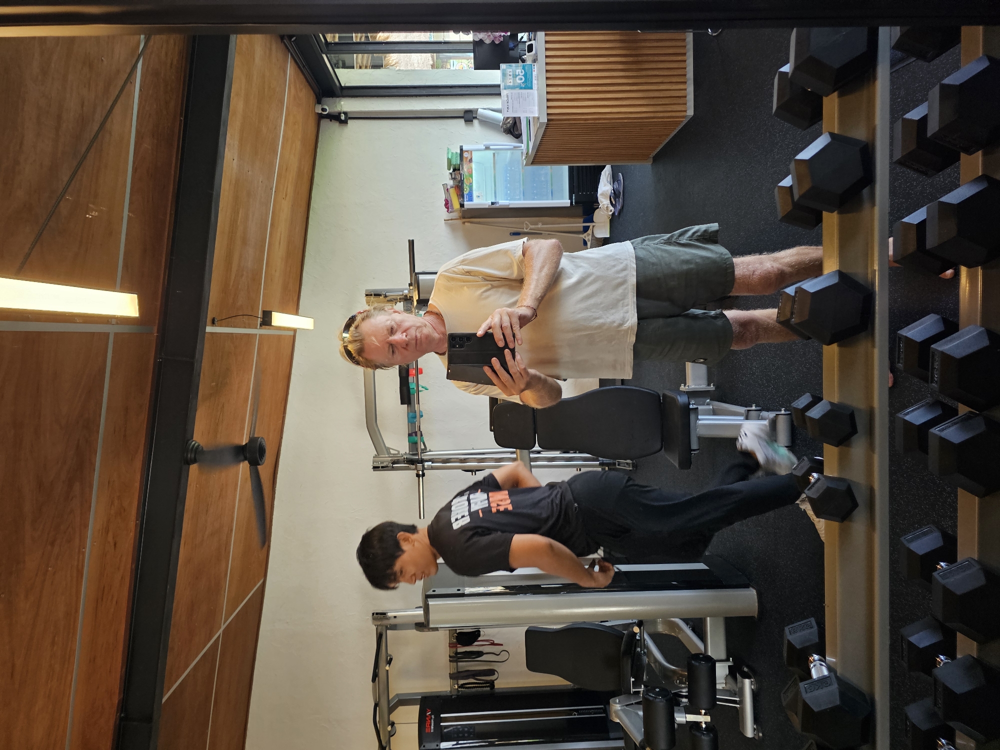 In Denpasar, Indonesia, a man with blonde hair and sunglasses is taking a mirror selfie in a gym. He is wearing a light-colored t-shirt and olive-green shorts and holding a black smartphone. To his left, a younger man wearing a black t-shirt with orange lettering and black pants is walking away from the camera. The gym is equipped with various weight machines and a rack of dumbbells is visible in the foreground. The ceiling is made of wood panels with exposed beams, and a ceiling fan is spinning. There is also a reception desk with a computer and a refrigerator in the background.