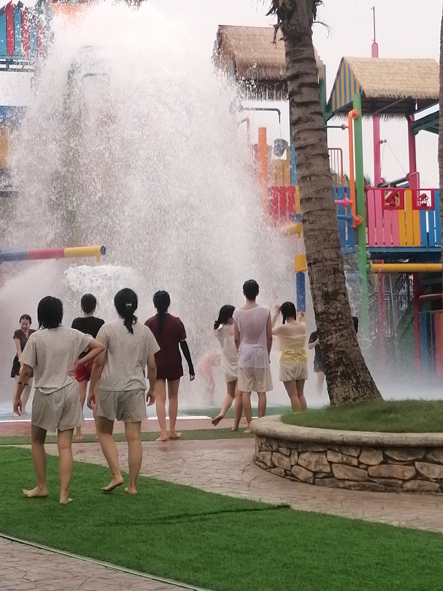 The image depicts an active scene at an outdoor water park, likely in Bai Chay Ward, Vietnam. Several people, primarily adults and mostly viewed from behind, are gathered in a paved area, intentionally engaging with a large cascade of water. A massive volume of water is gushing downwards from an elevated structure, creating a dense, white curtain that envelops the participants.
The individuals are dressed in light, casual clothing like t-shirts and shorts, which appear wet from the water. They are either standing directly in the path of the spray or walking towards/away from it, indicating they are enjoying the water feature. One woman on the far left is partially visible and appears to be smiling.
The water park environment features colorful structures with platforms, railings, and playful elements. Some of these structures are topped with thatched roofs, contributing to a tropical or resort-like ambiance. Two prominent palm trees frame the scene on the right, reinforcing this tropical feel. The ground consists of patterned paving stones, with sections of artificial green turf and a low, stone-faced planter visible in the foreground.
The time of day appears to be daytime, possibly overcast, as the lighting is soft with no harsh shadows. There is no visible text in the image. The overall impression is one of leisure and enjoyment at an aquatic recreational facility.