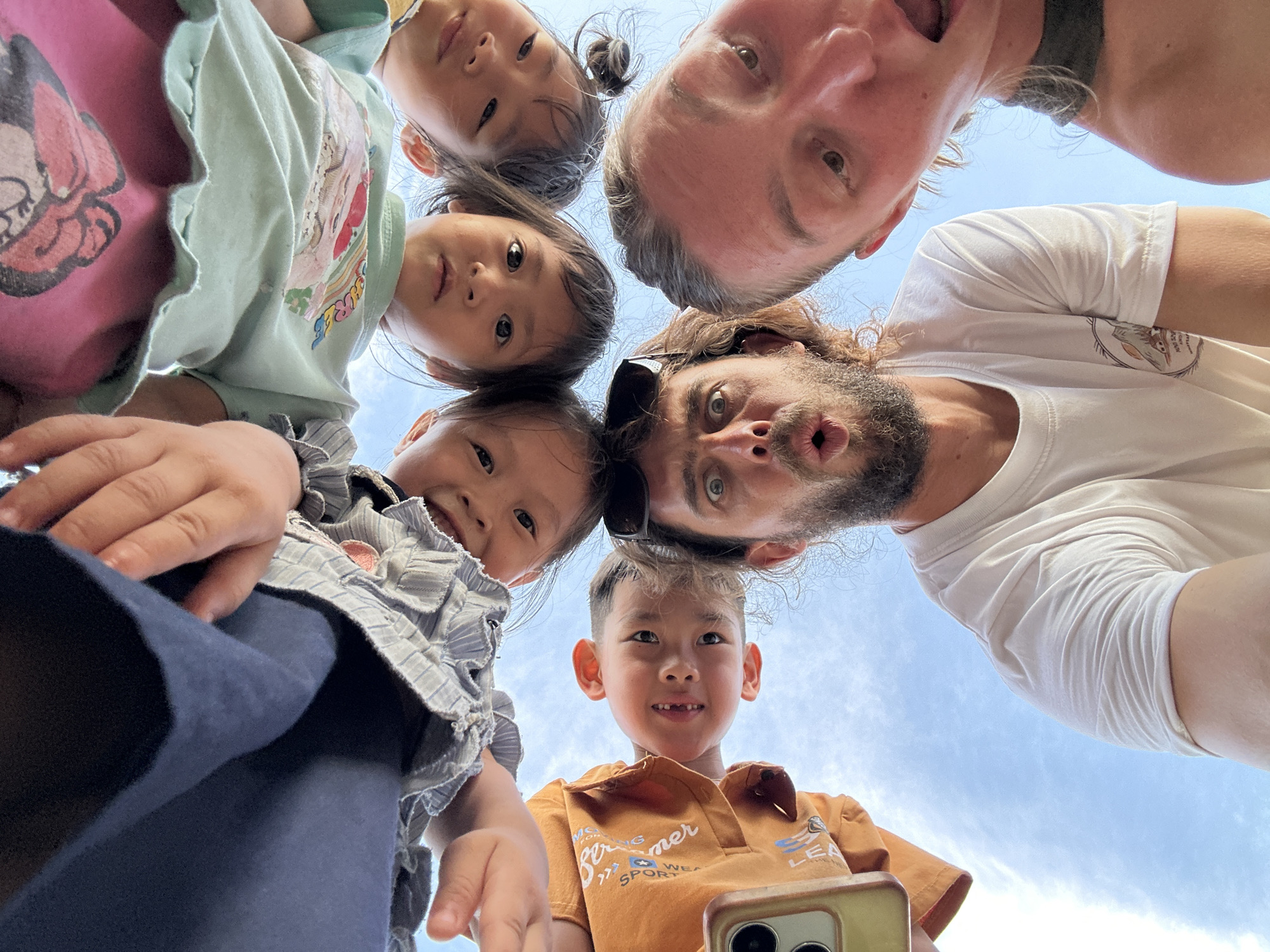 This image is a low-angle upward-facing group portrait, described as being taken in Gia Hội, Vietnam. Six individuals are prominently featured, looking directly down into the camera lens, forming a semi-circular arrangement against a clear blue sky.
At the bottom-center is an adult male with a dark beard and curly hair, wearing sunglasses on his head and a white t-shirt; his eyes are wide, and his mouth is open in an "O" shape. To his immediate right, a second adult male with lighter hair is partially visible, with an open mouth and a surprised expression.
Above these adults, four children are positioned. On the left, a child in an orange t-shirt smiles towards the camera. Adjacent to this child, in the bottom-left corner of the frame, is a tan-colored smartphone, partially held by a hand, showing multiple camera lenses. Moving right, a child in a light green t-shirt holds a neutral expression. To their right, a child in a pink t-shirt also maintains a neutral expression. Further to the left and slightly above the orange-shirted child, a fourth child in a light blue striped top with ruffled detailing is partially visible, with hands reaching downwards into the frame.
The background consists entirely of a bright, clear blue sky with minimal cloud formations, indicating an outdoor setting with ample natural light.