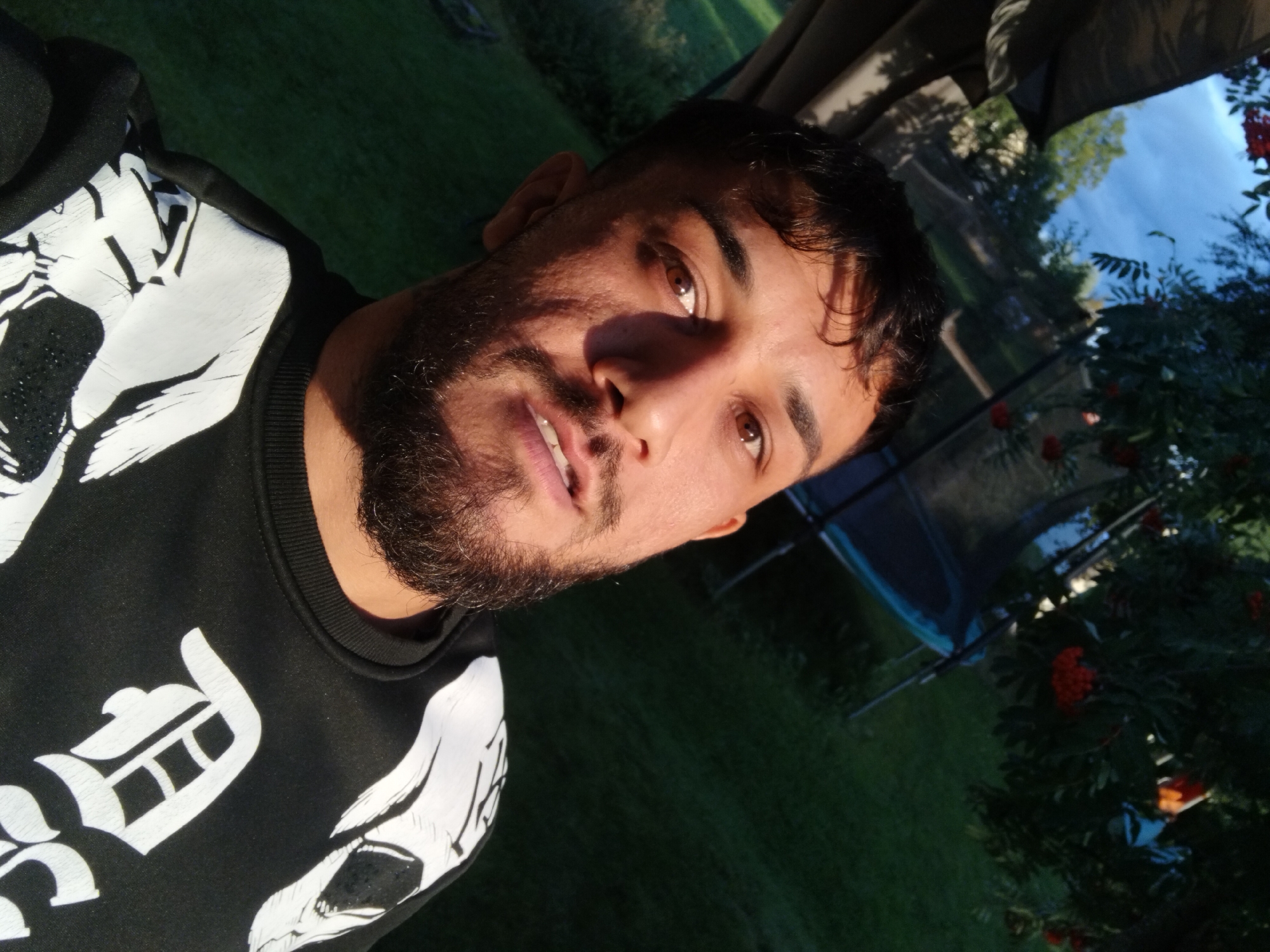 A close-up, selfie-style portrait of a man with dark hair, a beard, and brown eyes. He is wearing a black sweatshirt with white graphic print. The man is outdoors, with a background of green grass, trees with red berries, and a blue sky. A trampoline is partially visible in the background. The lighting suggests it is either early morning or late afternoon. The location is identified as Rätan, Sweden.