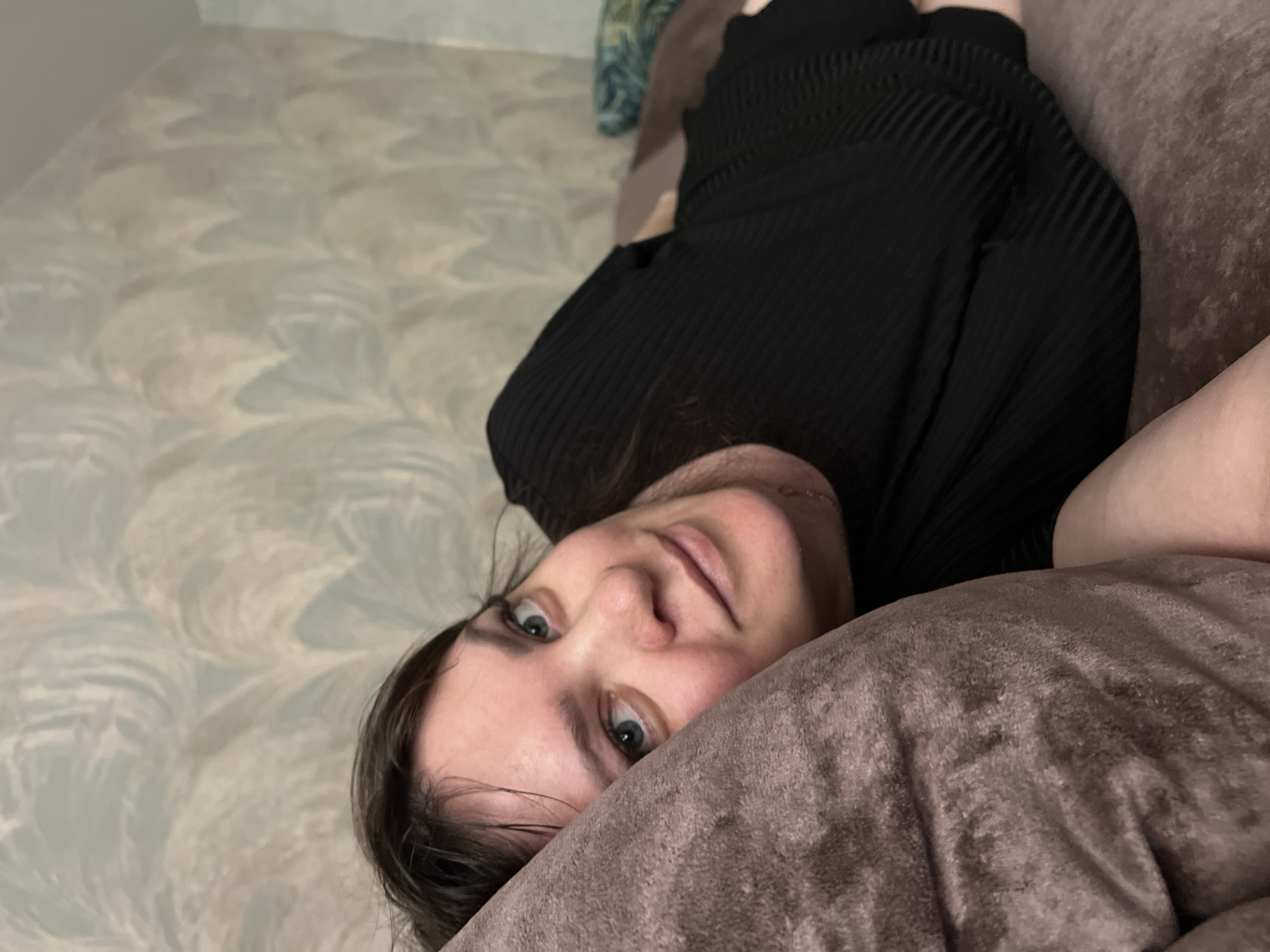 A woman with light skin and blue eyes is lying down on a brown, upholstered surface, likely a sofa or bed. She is wearing a black, ribbed top. A decorative wallpaper can be seen in the background. The location information provided, Noviy, Russia, is not discernible from the image.