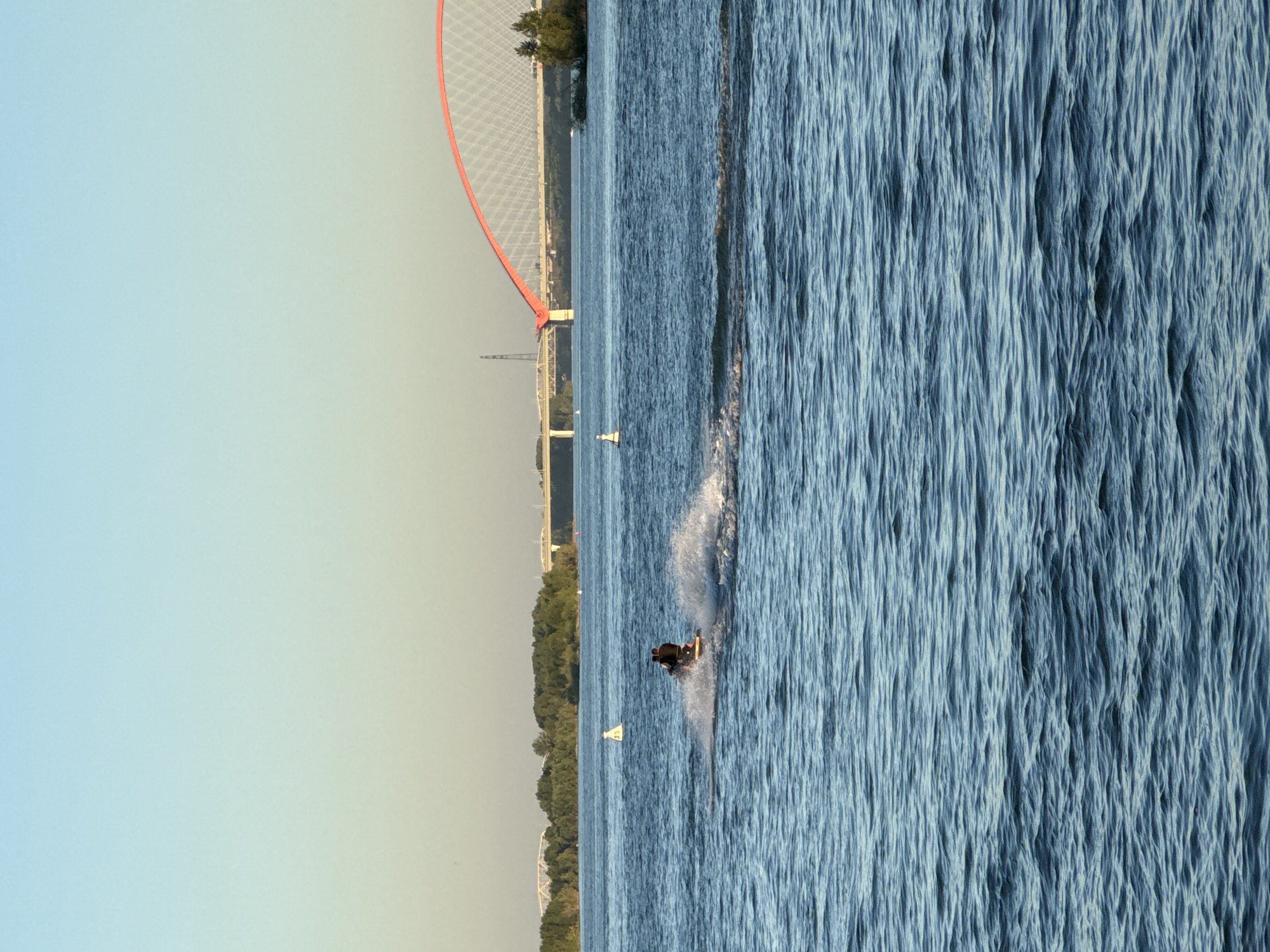 The image shows a scenic view of the Ob River in Novosibirsk, Russia. The focus is on a jet ski with two people on it, creating a wake as they speed across the water. In the background, a large, red-arched bridge spans the river, a prominent landmark of the city. The sky is clear and blue, indicating pleasant weather, with a subtle gradient from light blue at the top to a softer yellow-beige hue near the horizon, possibly due to the time of day, suggesting it might be sunset or sunrise. The water is a deep blue, reflecting the sky and creating a dynamic texture with ripples and waves. Several buoys are visible in the water, marking channels or hazards. The distant shoreline is lined with lush green trees, adding to the natural beauty of the scene.