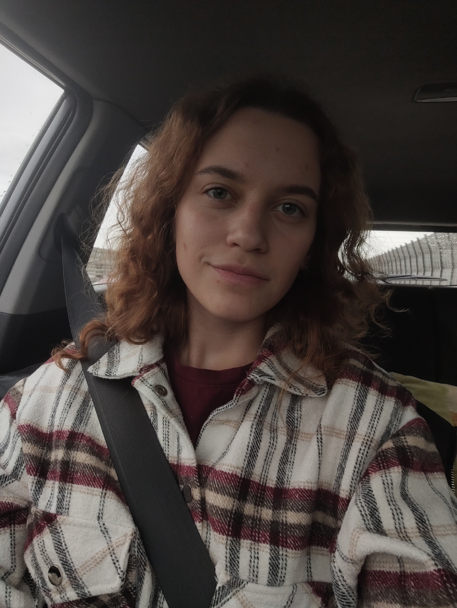 A young woman is seated in the interior of a car, looking directly at the camera with a neutral, subtly smiling expression. She has medium-length, wavy reddish-brown hair and light-colored eyes. She is wearing a plaid button-up jacket with patterns in shades of white, cream, dark red, and black, over a dark red top. A black seatbelt crosses her torso diagonally from her left shoulder.
Through the car windows, an overcast sky is visible, with water droplets on the glass indicating rain or recent precipitation. The blurred background outside the car indicates daylight. Elements of the car's interior, including the headliner, door frame, and rearview mirror (visible in additional crops), are apparent. The soft, diffused lighting suggests an overcast daytime setting. No text is visible in the image.