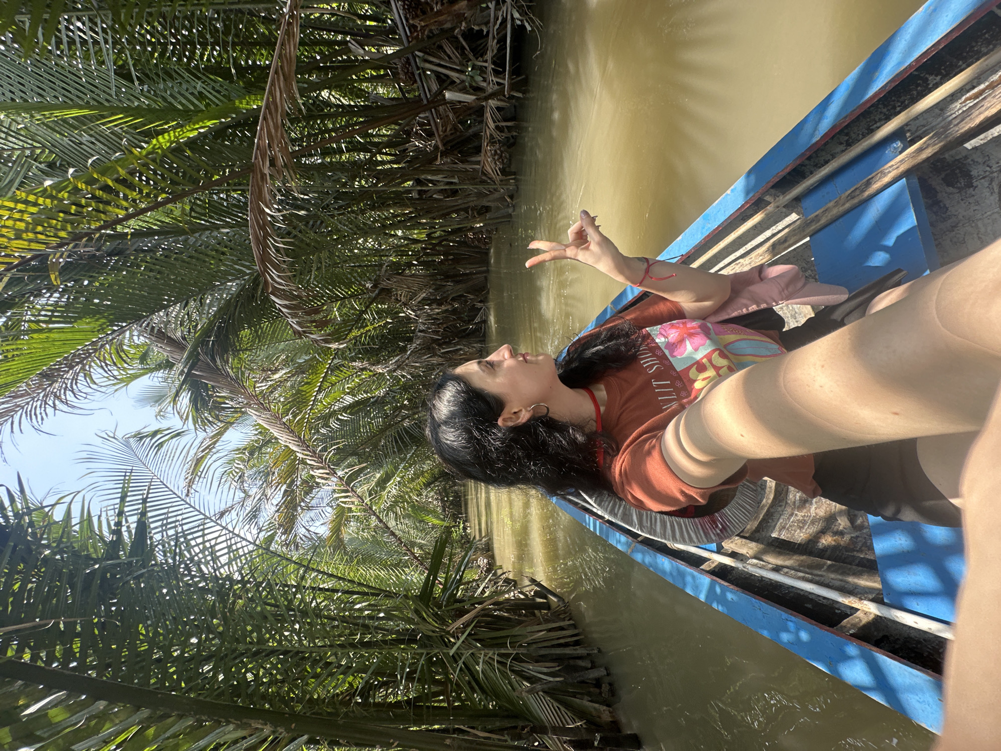 A person is in a boat floating on a river surrounded by dense, green vegetation, potentially located in Tân Thạch, Vietnam. The water is murky and brown. The boat is blue. The person has dark hair and is wearing an orange t-shirt. The person is extending fingers in the air.