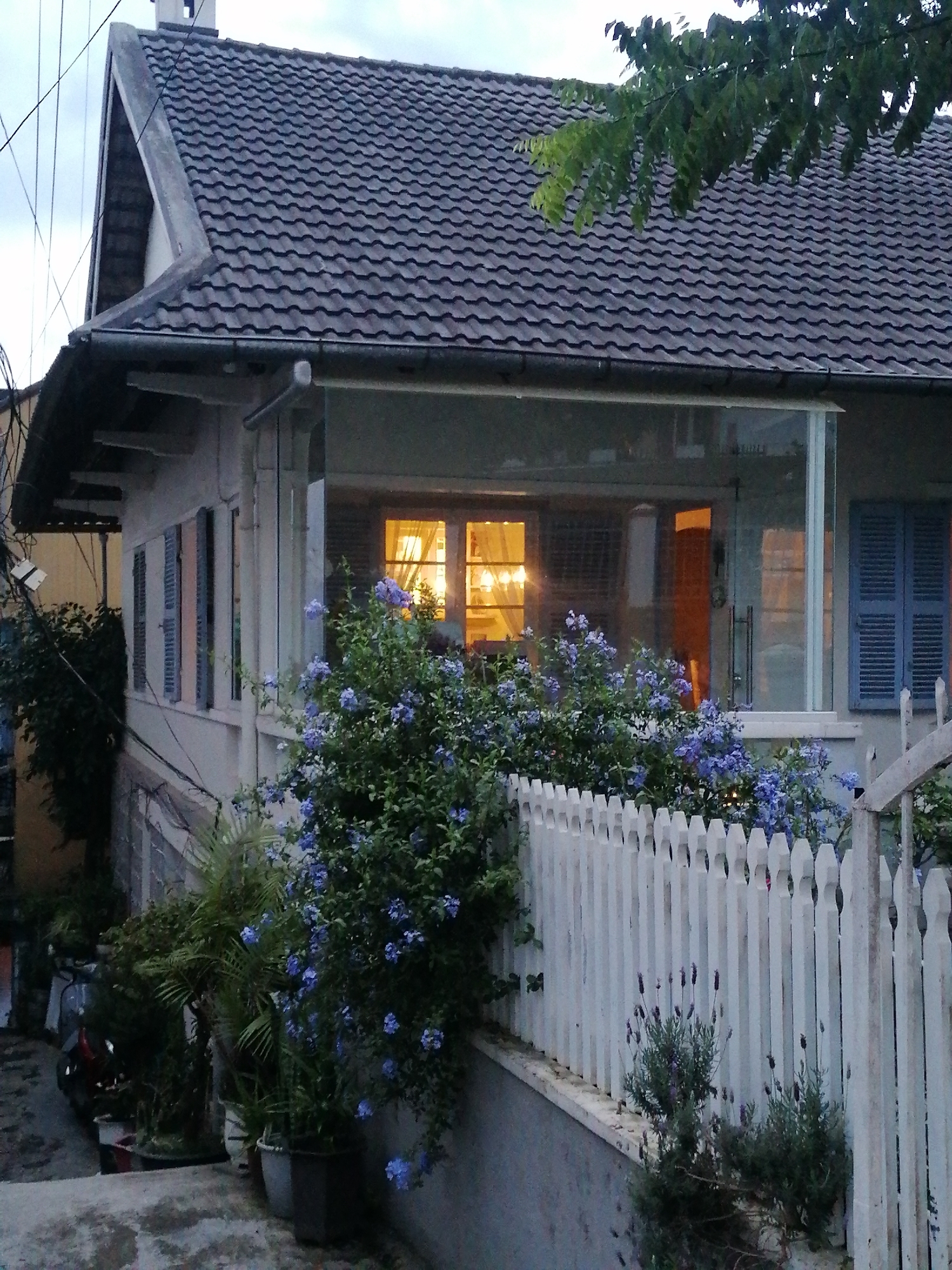 A wide shot captures a white, two-story house with a dark tiled roof on a cloudy day. The house features a glass-enclosed porch on the first floor, illuminated from within with warm light. Blue shutters are visible on the windows. In the foreground, a white picket fence encloses a garden with blooming purple flowers and lush green foliage. Several potted plants and a parked scooter are also visible to the left. The image appears to be taken in Cam Ly, Đà Lạt, Vietnam, given the architectural style and vegetation typical of the region.