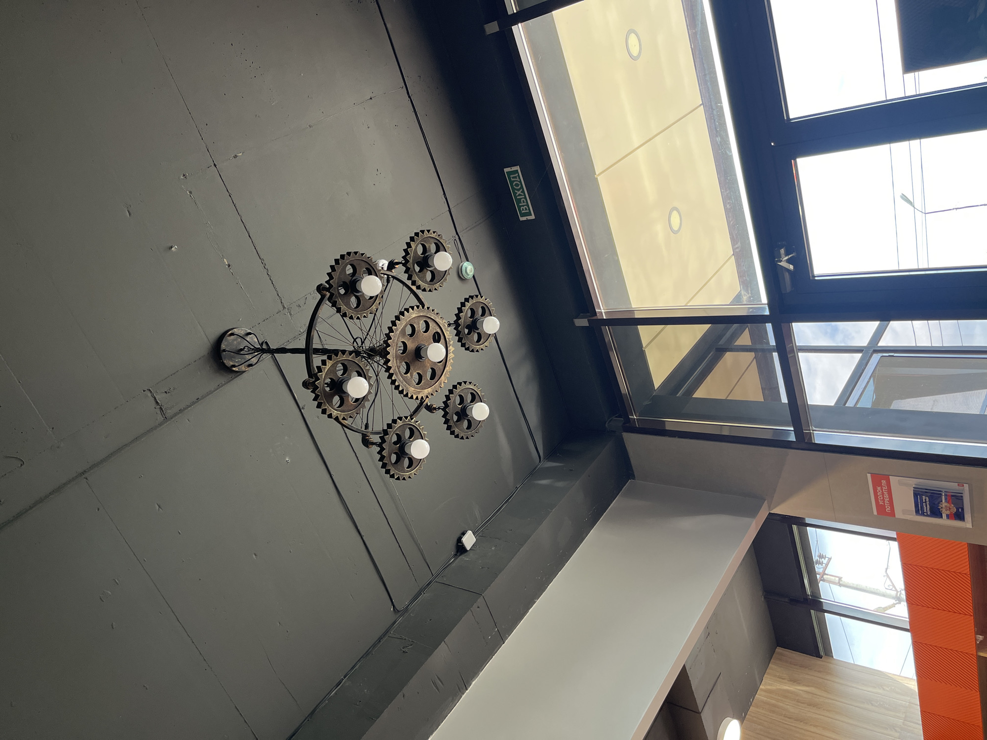 The image shows an interior scene in Kaspiysk, Russia. The view is directed upward, focusing on the ceiling and upper parts of the interior. The ceiling is dark, possibly gray or black, and features a decorative light fixture resembling a bicycle wheel with gears and light bulbs. A green sign with white lettering, possibly indicating an exit, is visible near the edge of a skylight or window structure. The skylight is covered with semi-transparent glass, allowing some natural light to enter. The interior architecture includes gray and white painted walls, with a sign in Russian on the wall and an orange decorative element. The environment appears to be inside a commercial space or public building.