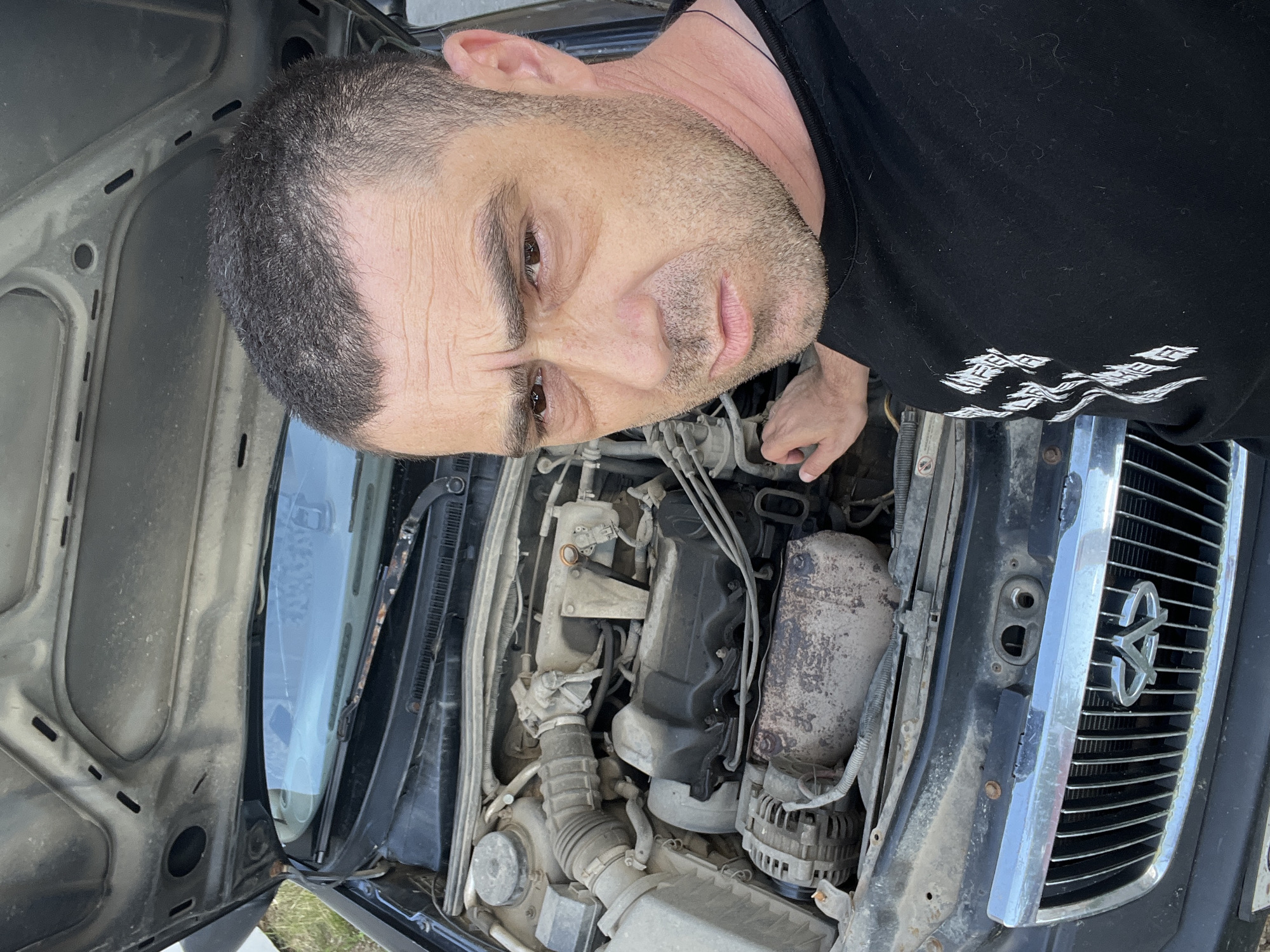 A man is taking a selfie with the hood of a car open, revealing the engine. The man is in the foreground, looking at the camera with a concerned expression. He is wearing a black t-shirt. The car's engine compartment is dusty and appears to be in need of maintenance. The car's grille is visible at the bottom of the image, showing the Chery logo. The background is blurred and appears to be an outdoor setting.