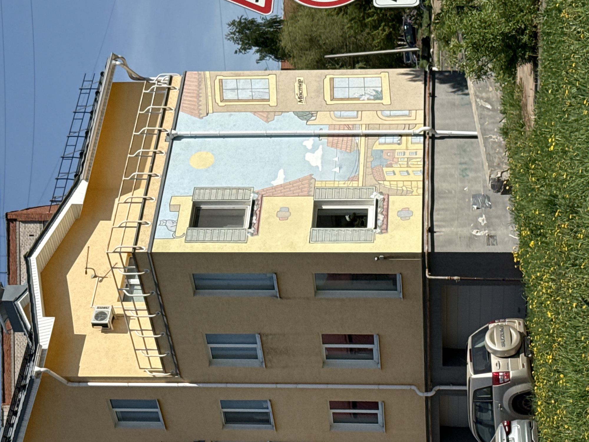 The image shows a building with a mural in Votkinsk, Russia. The building is light yellow with a ladder on the side. The mural depicts a European cityscape with windows and a blue sky. Below the mural, the building has several rectangular windows. A silver SUV is parked next to the building, partially obscured by green foliage. The sky is blue. Traffic signs are visible in the background.