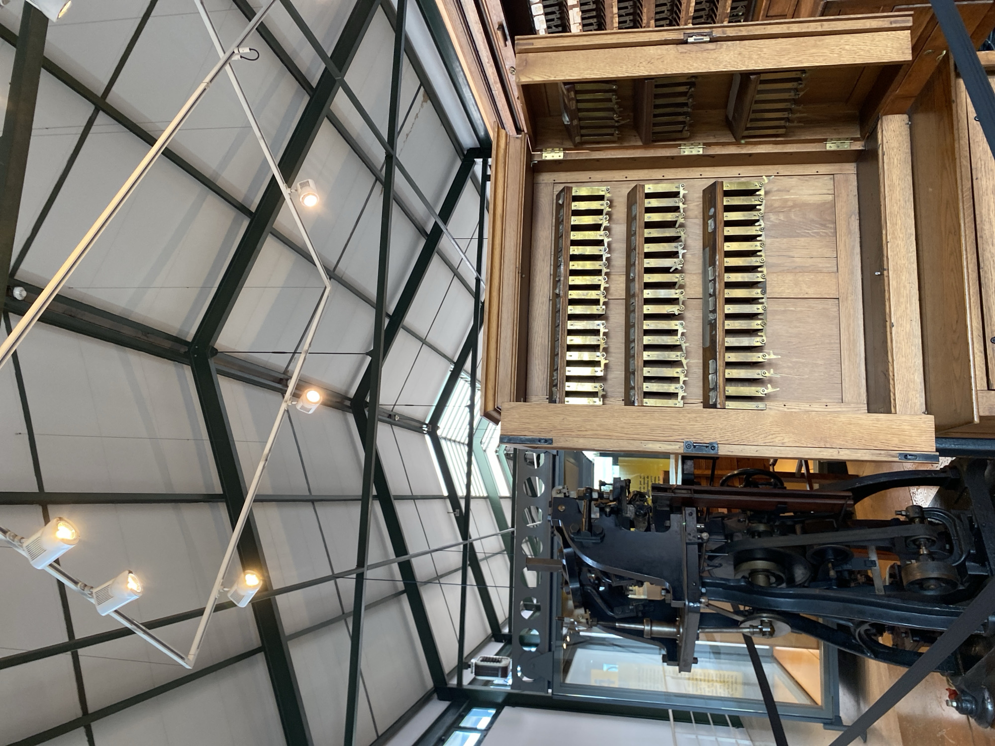 The image shows an interior view with a complex geometric ceiling structure of metal beams and white panels, illuminated by several spotlights. To the right is an old, wooden typesetting machine with rows of metal letters, standing on a complex black metal framework above a glass case. The location appears to be the Museo Casa Natal de Jovellanos in Gijón/Xixón, Spain, as the museum houses an old typesetting machine and has a distinctive ceiling structure. No people are visible.