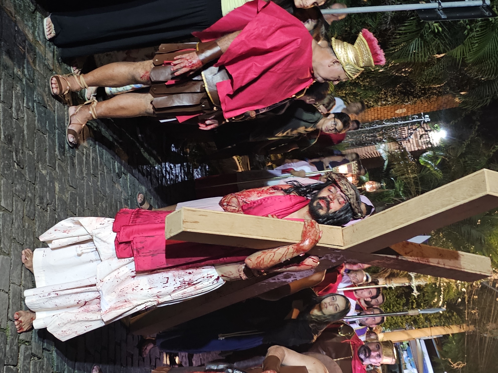A religious procession is taking place in the city of Armação dos Búzios, Brazil, portraying the crucifixion of Jesus Christ. A man with a beard and long hair, representing Jesus, lies prone, carrying a wooden cross. His clothing is white and red, stained with fake blood. He is surrounded by people dressed as Roman soldiers and other biblical figures. A soldier with a golden headdress and a red cape stands nearby, his arms covered in fake blood. The procession is occurring on a cobblestone street. There are bystanders watching the procession.